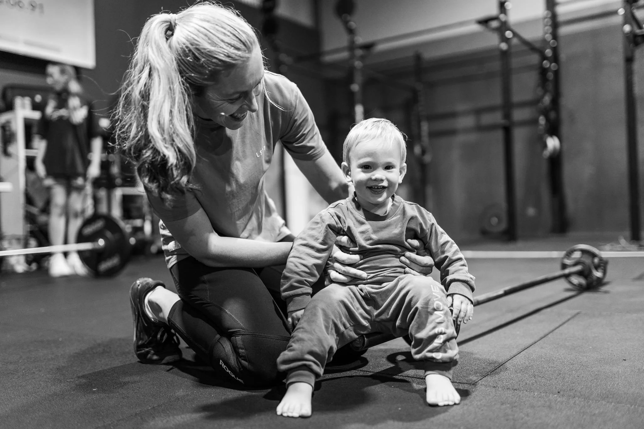 A woman and a young boy with Down syndrome are smiling while sitting on a gym floor. The woman is leaning over the boy, who is sitting with his legs crossed, in a gym with workout equipment in the background.