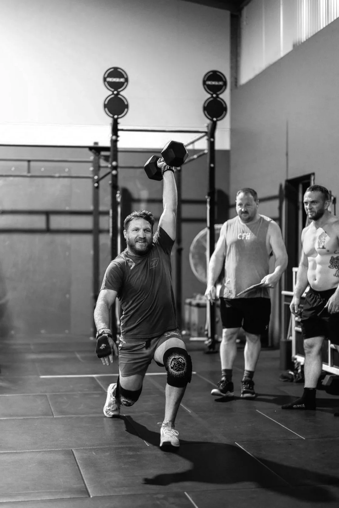 A man with a beard participating in CrossFit, lifting a dumbbell overhead in a gym. Two men stand nearby watching; one is shirtless and the other in a tank top. Gym equipment and wall-mounted rings are visible in the background.