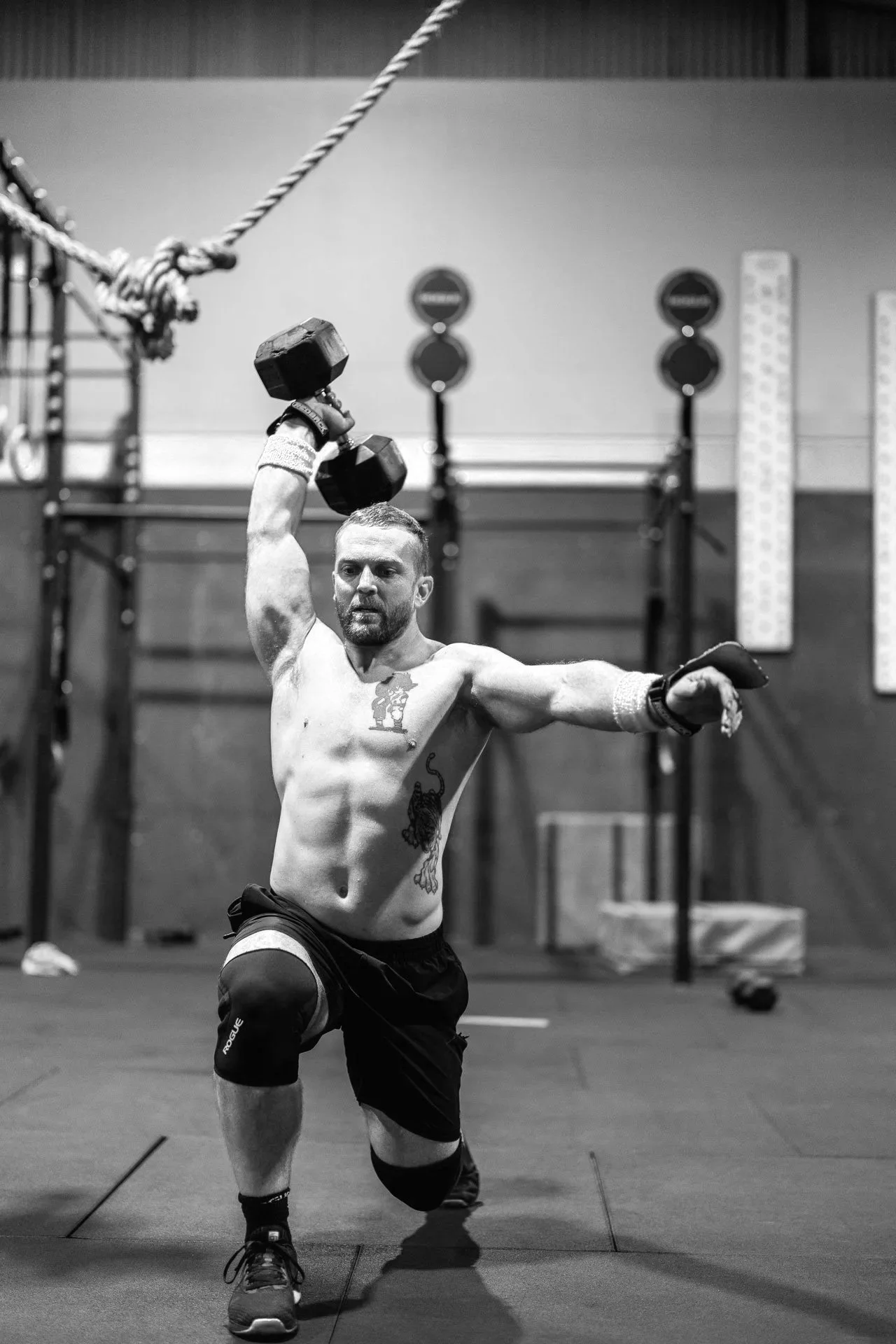 A shirtless man with tattoos on his chest and arm, wearing shorts, kneeling on one knee in a gym, lifting a dumbbell above his head with one hand, with gym equipment and weights in the background.