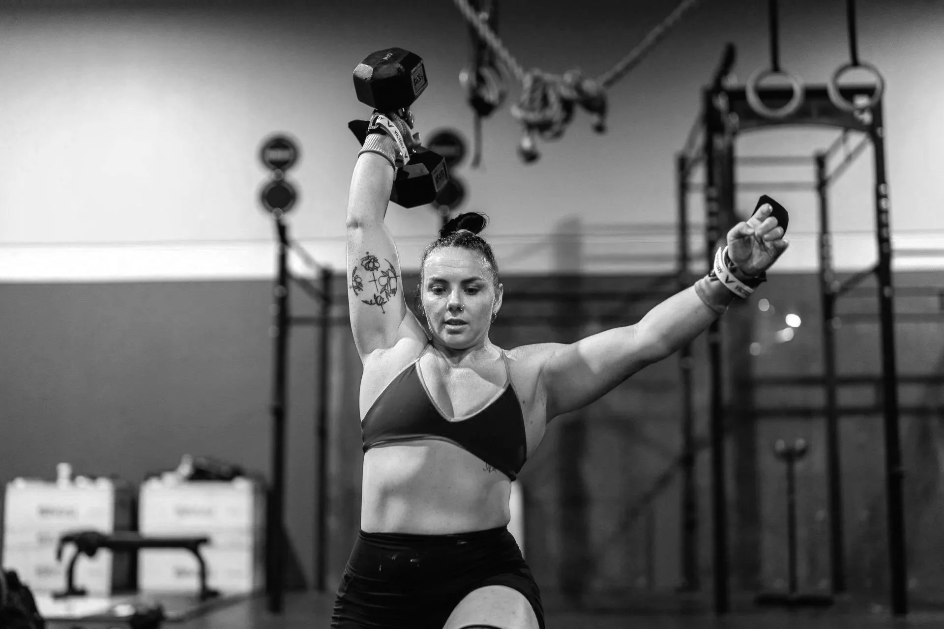 A woman lifting a dumbbell in a gym, with gym equipment and gymnastic rings in the background, captured in black and white.