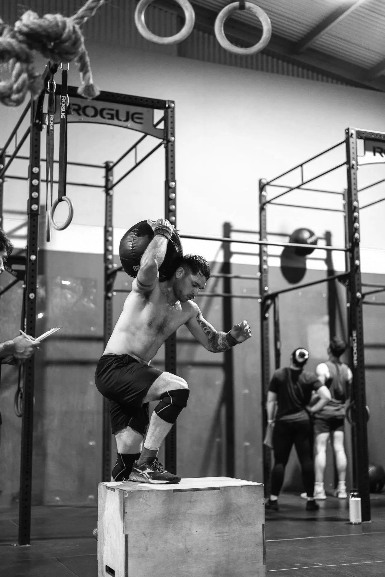 A man performing a box jump exercise with a medicine ball in a gym, with gymnastic rings and workout equipment in the background.
