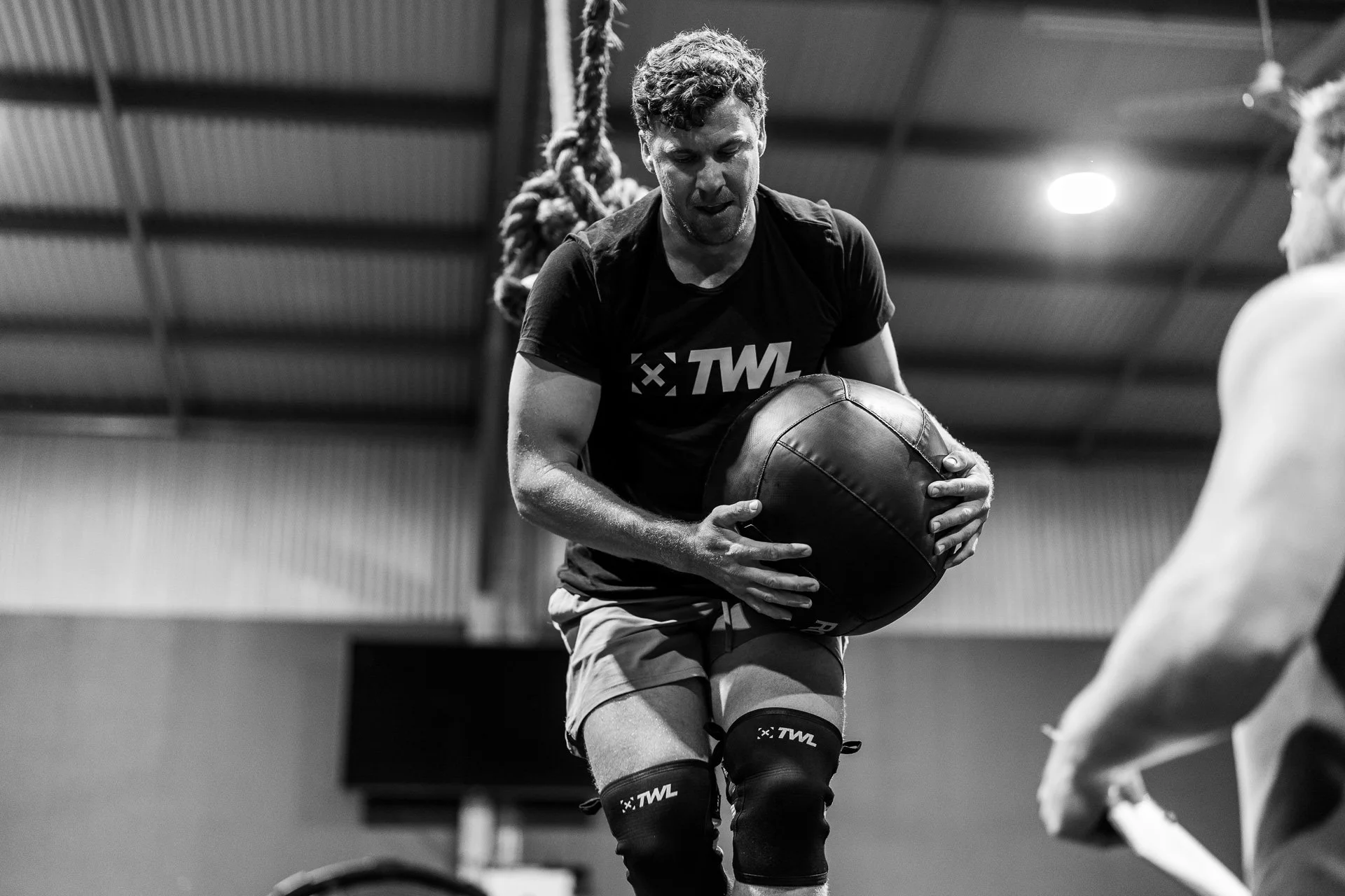A man preparing to throw a medicine ball during an indoor workout session, with a trainer in front of him.