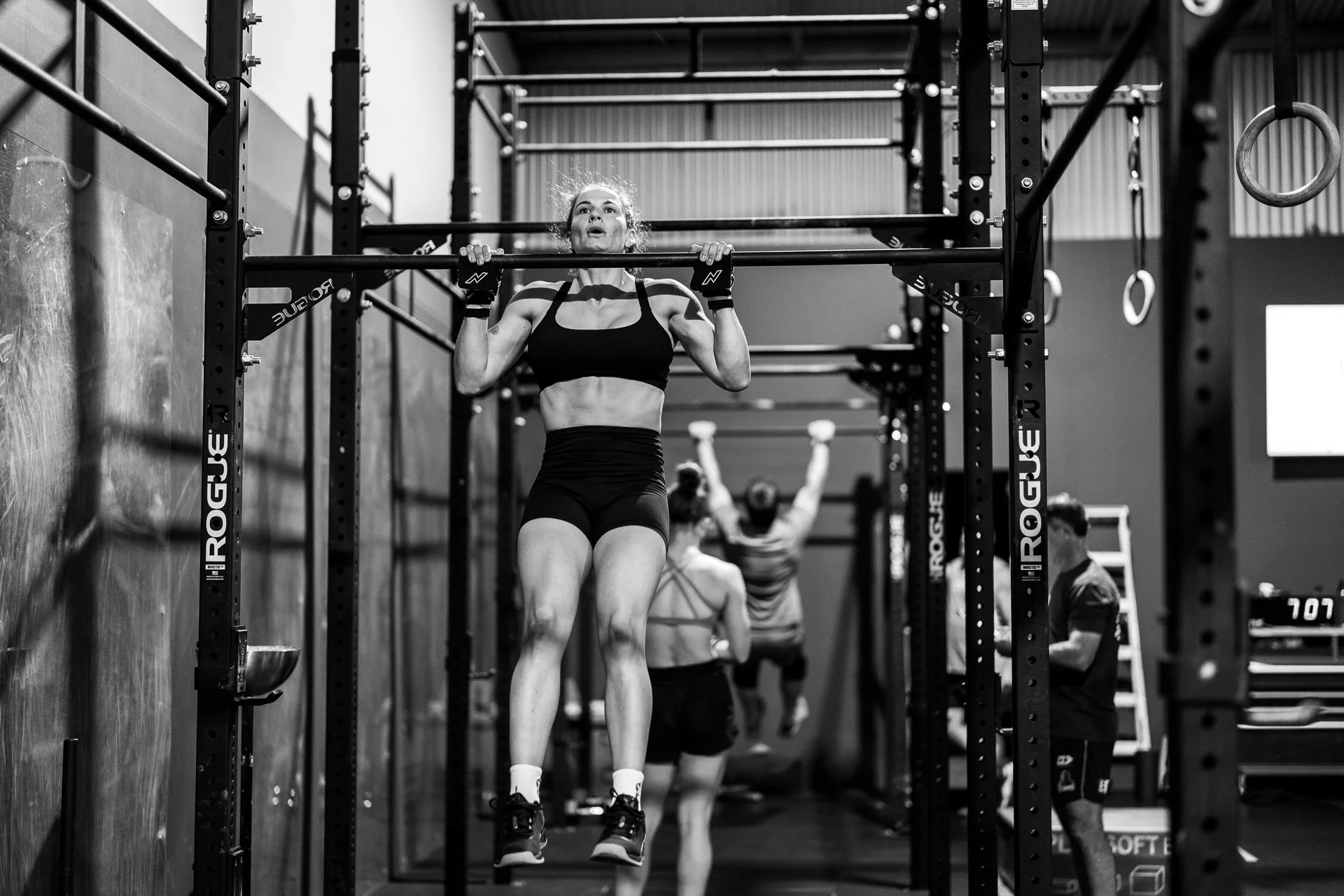 A woman in athletic attire performing a pull-up in a gym, with other individuals working out in the background.