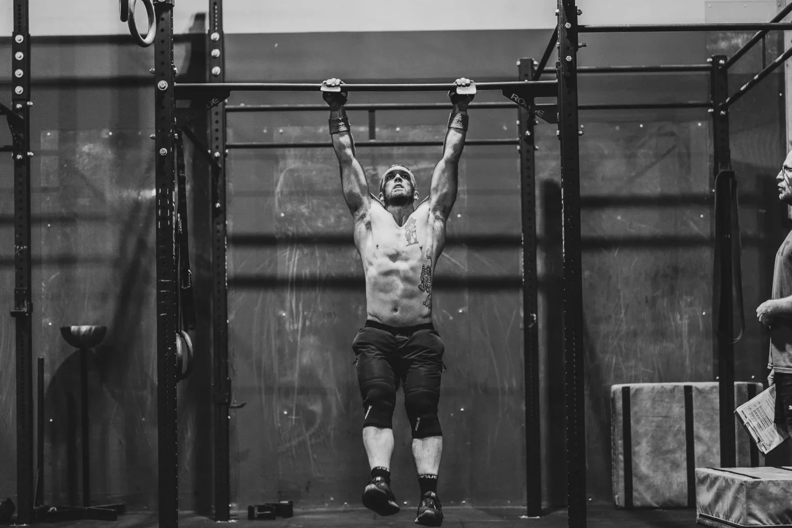 A shirtless man with tattoos doing a pull-up exercise on a pull-up bar in a gym. Another person is standing nearby holding a paper or notebook.