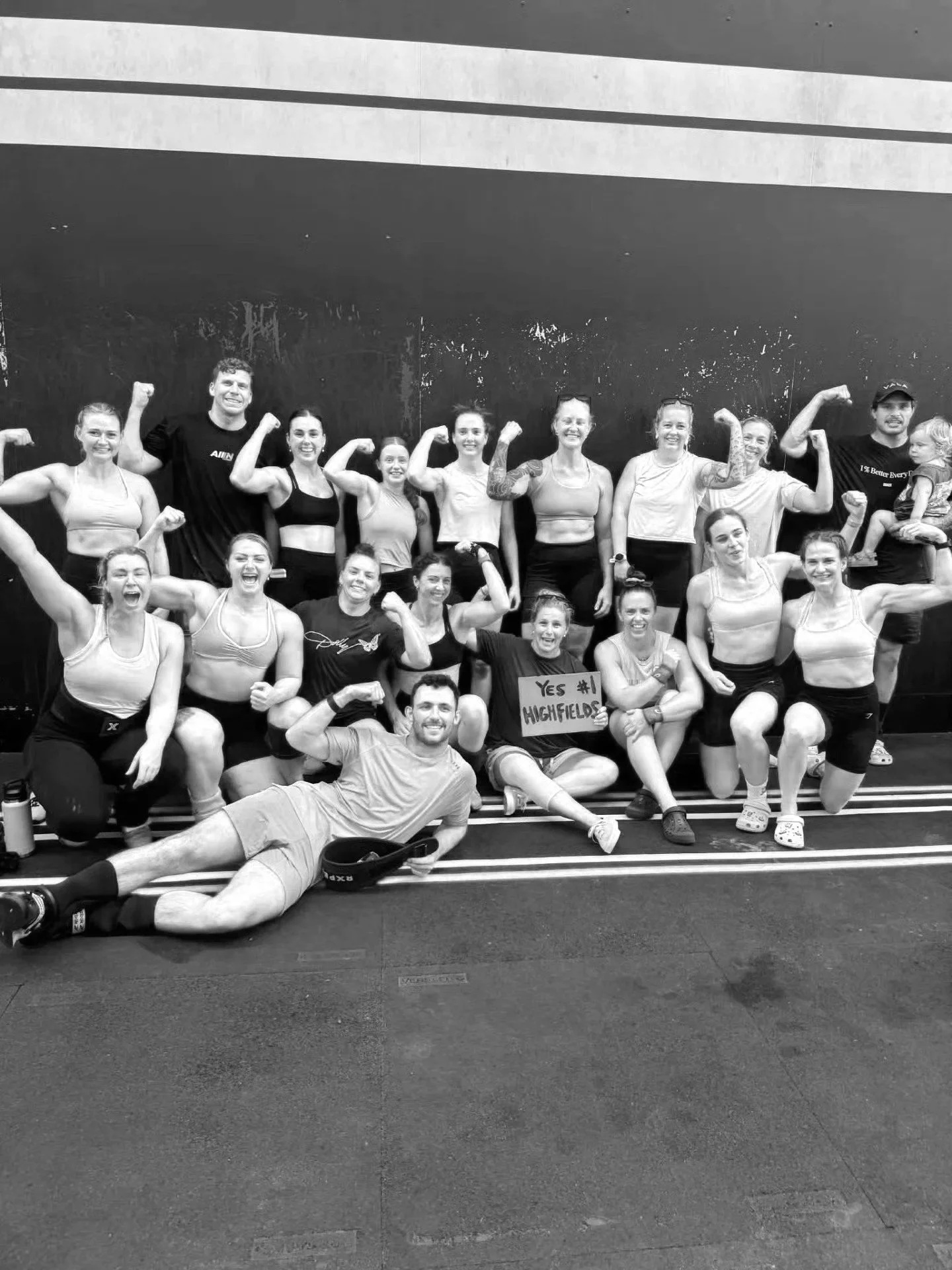 A group of people in sportswear flexing their muscles in a gym or fitness center, with a mostly black wall in the background and a person lying in front of the group posing.