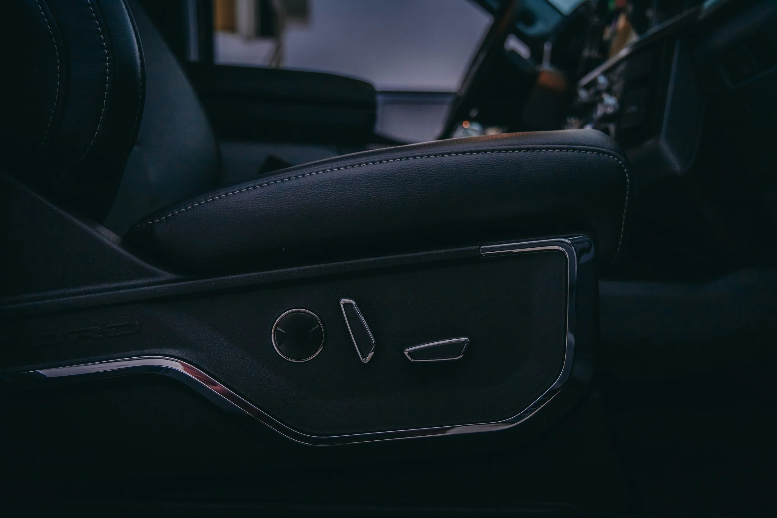Close-up of the interior of a luxury car, focusing on the black leather armrest with white stitching and the dashboard controls.