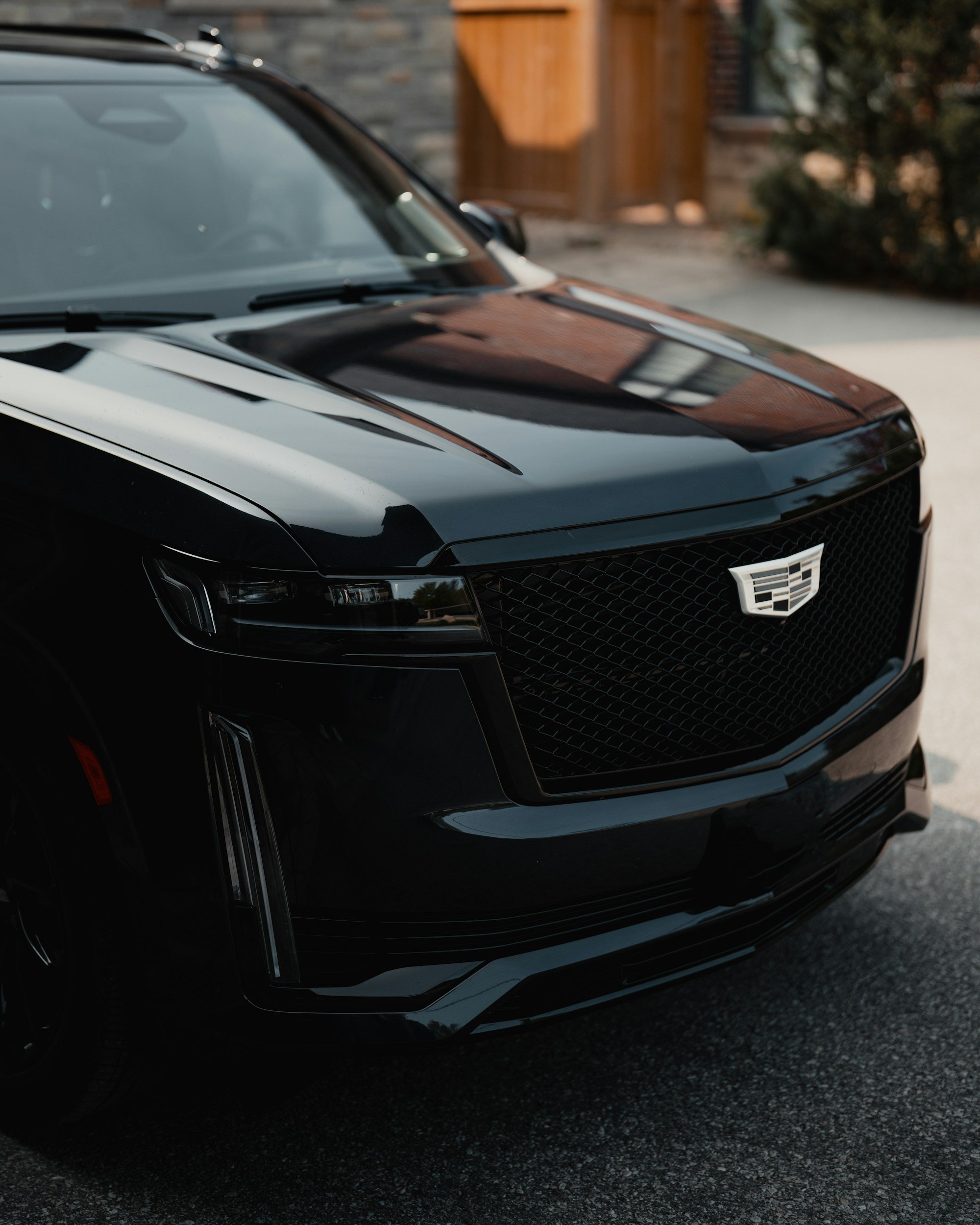 Front view of a black Cadillac car parked on a street with a wooden house and some bushes in the background.
