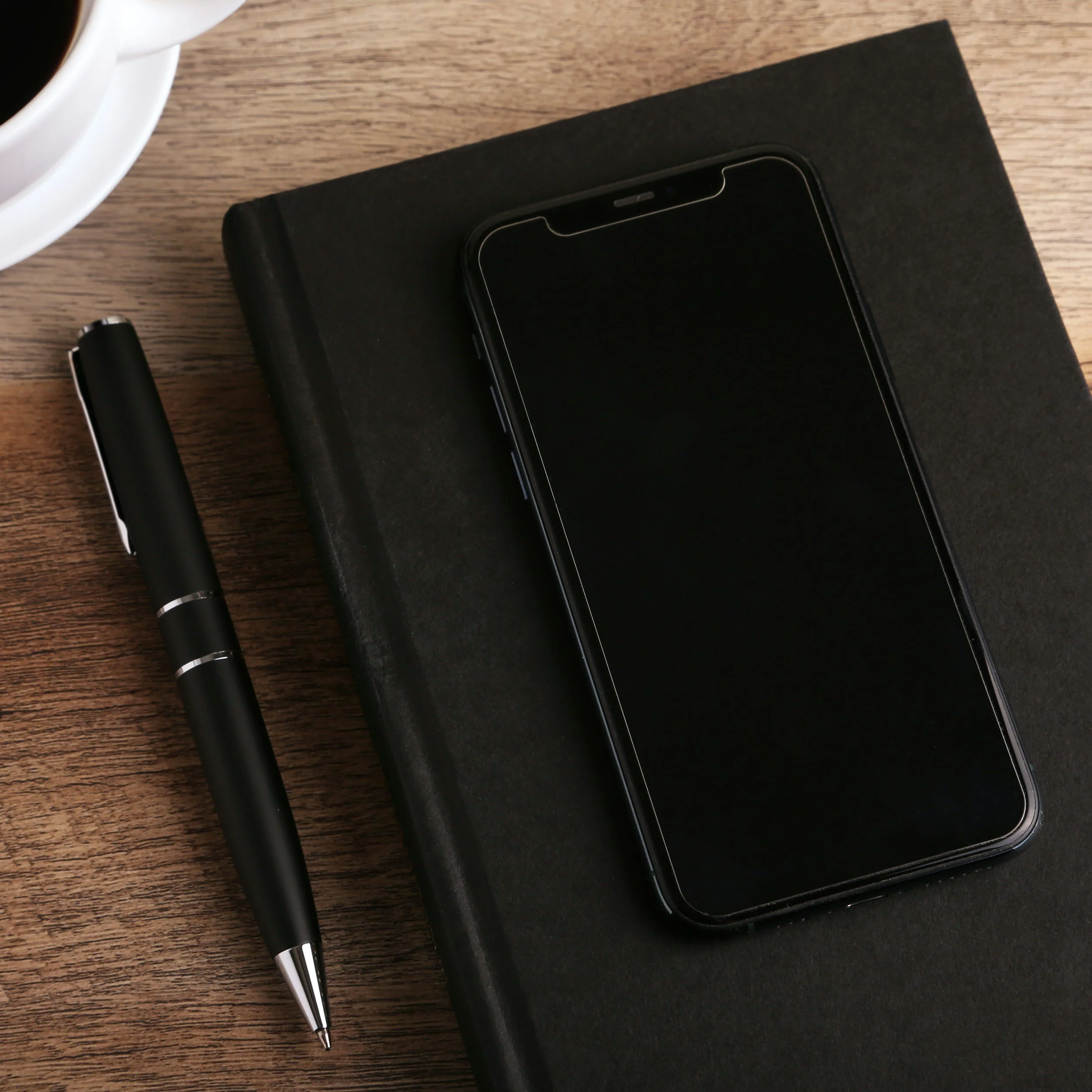 A top-down view of a wooden desk with a black smartphone, a black notebook, a black and silver pen, a black pencil, and a cup of black coffee in a white mug.
