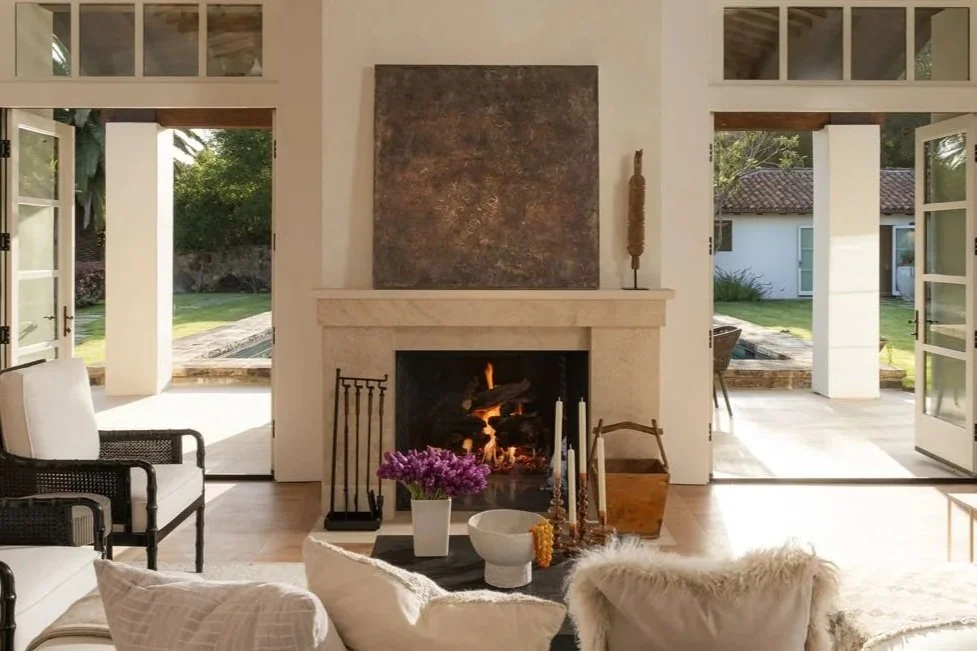 Living room with a fireplace, open doors leading outside to a yard, and furniture including armchairs and a coffee table with flowers and candles.