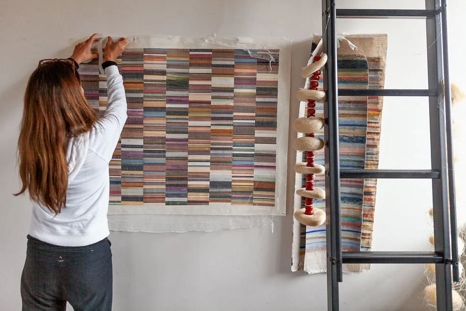 A woman hangs a colorful woven textile on a wall near a ladder with other textiles and decorative objects.