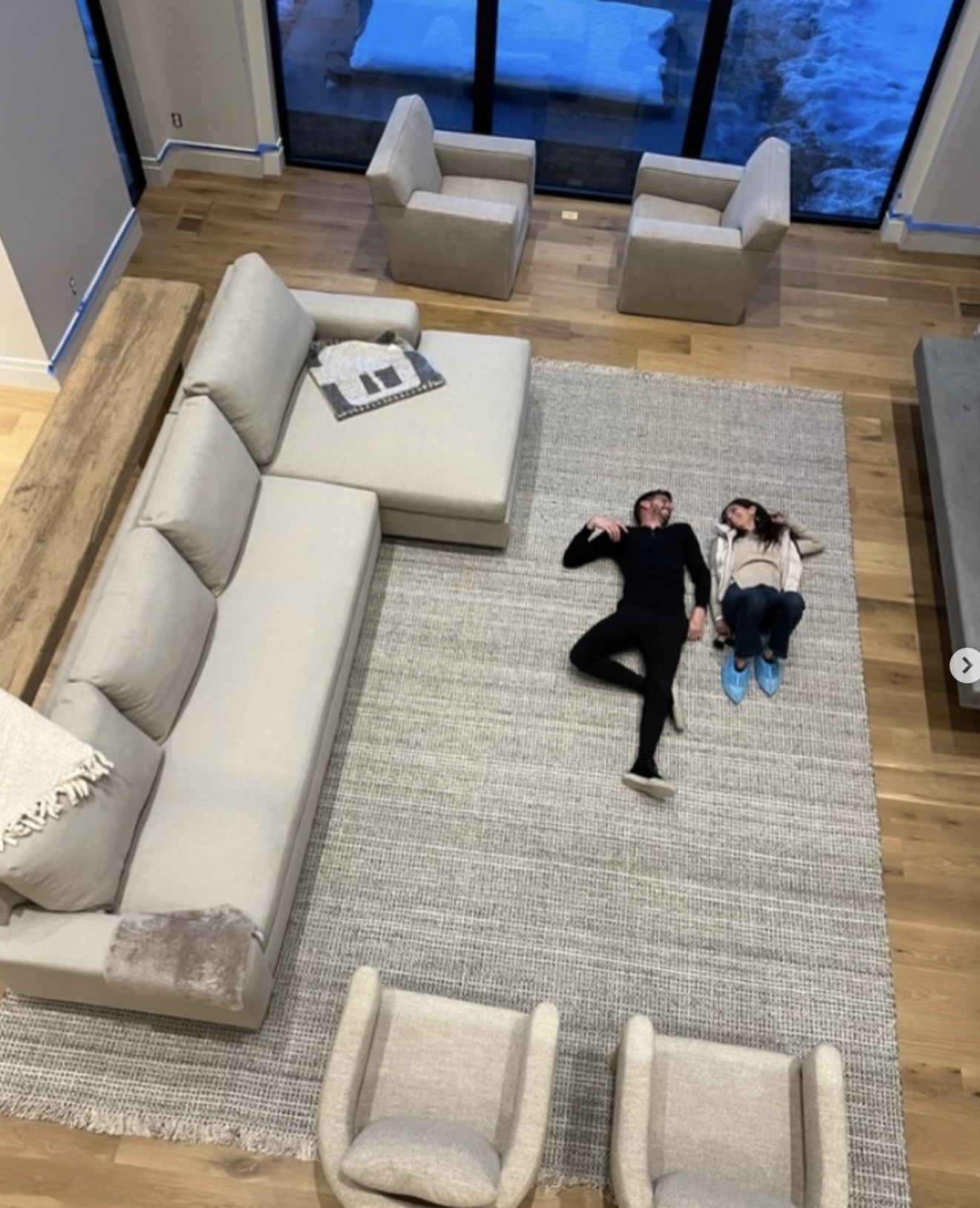 Two people lying on a rug in a living room, looking at each other and smiling. The room has a beige sectional sofa, two armchairs, a wooden side table, a large area rug, hardwood flooring, and a glass wall showing a snow-covered outdoor area.