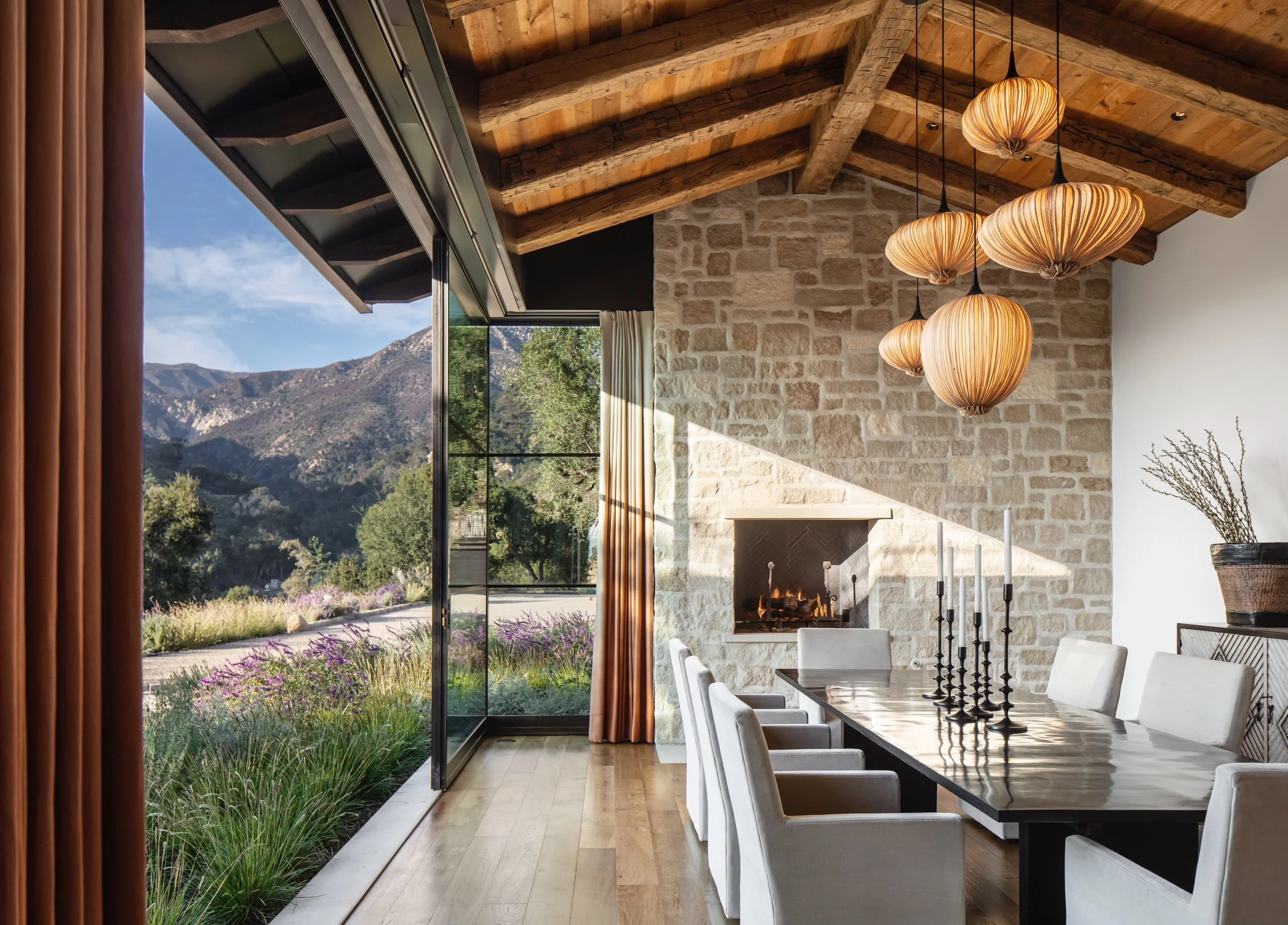 Interior of a dining room with a large window showing mountains and trees outside, wooden ceiling beams, hanging pendant lights, a stone fireplace, and a long table with white chairs.