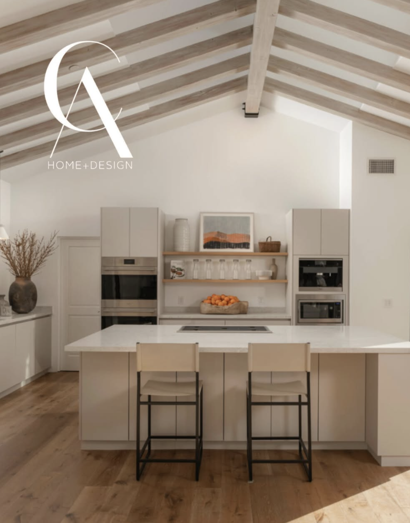 Modern white kitchen with a large island, open wooden shelves with glasses and a bowl of oranges, and built-in appliances. The ceiling features exposed wooden beams, and there is a decorative vase with dried plants on the side.
