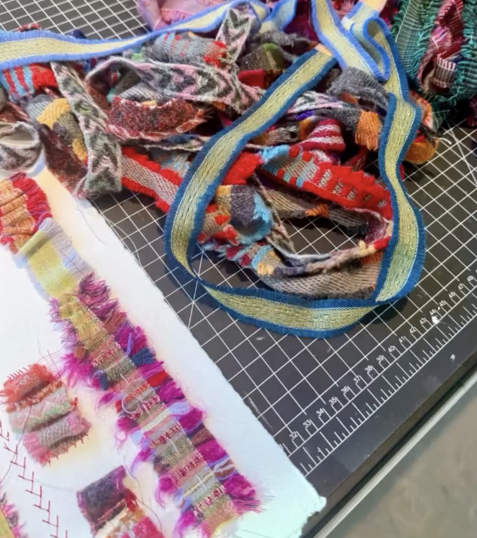 Colorful, patterned fabric scraps and woven ribbon on a cutting mat with a ruler, likely for crafting or textile art.