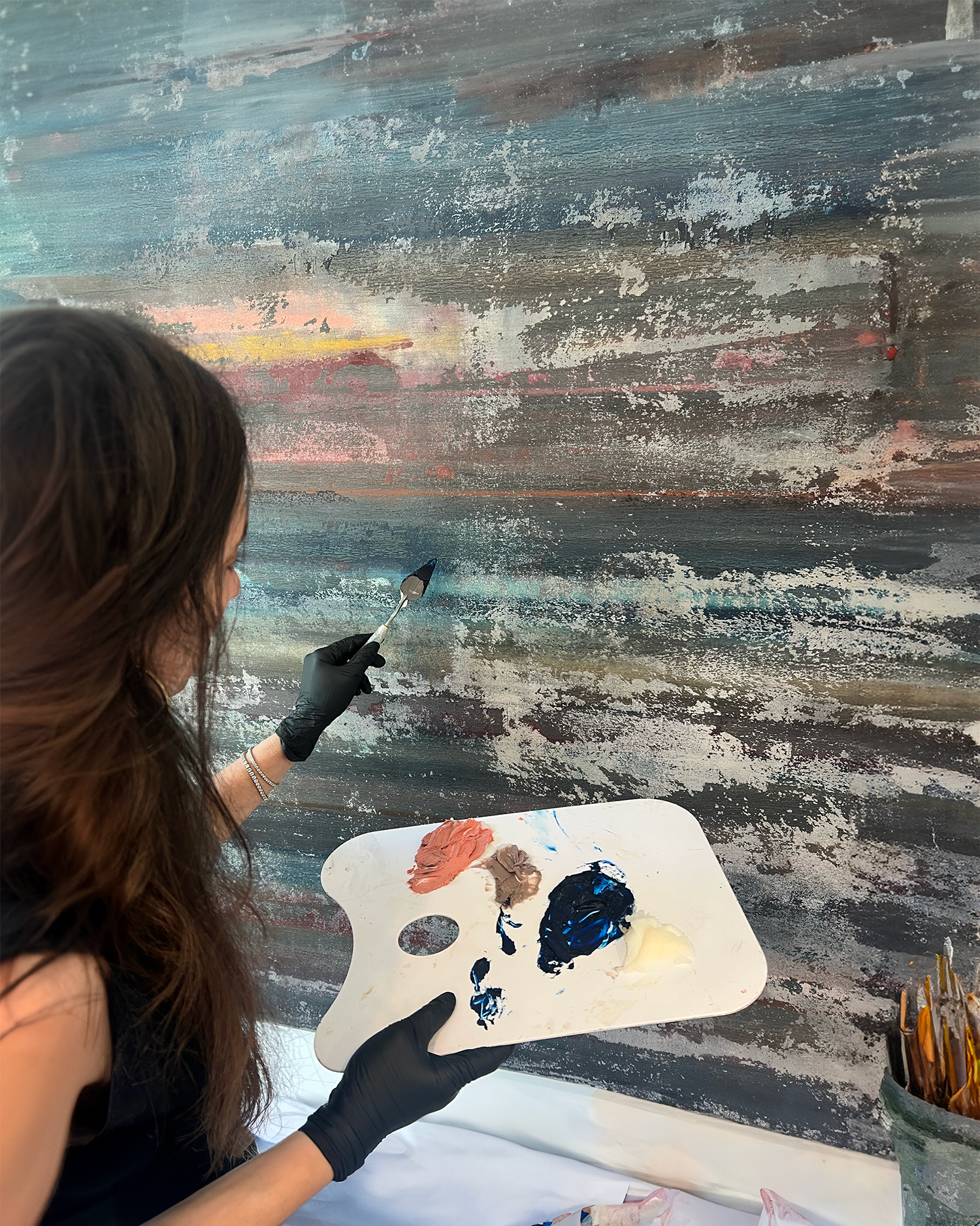 An artist wearing black gloves holding a painter's palette with different colors of paint, including blue and beige, working on an abstract landscape painting that depicts a sunset over water with a sailboat.