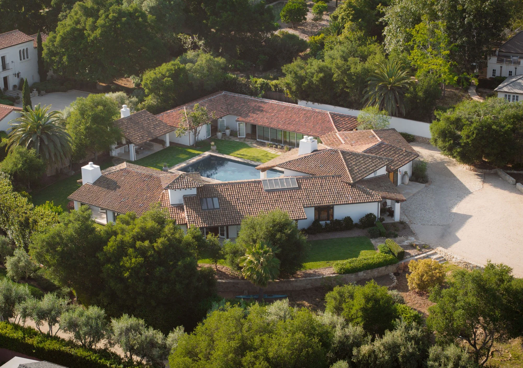 Aerial view of a residential property with a swimming pool, surrounded by lush green trees and white walls, with other houses visible in the background.