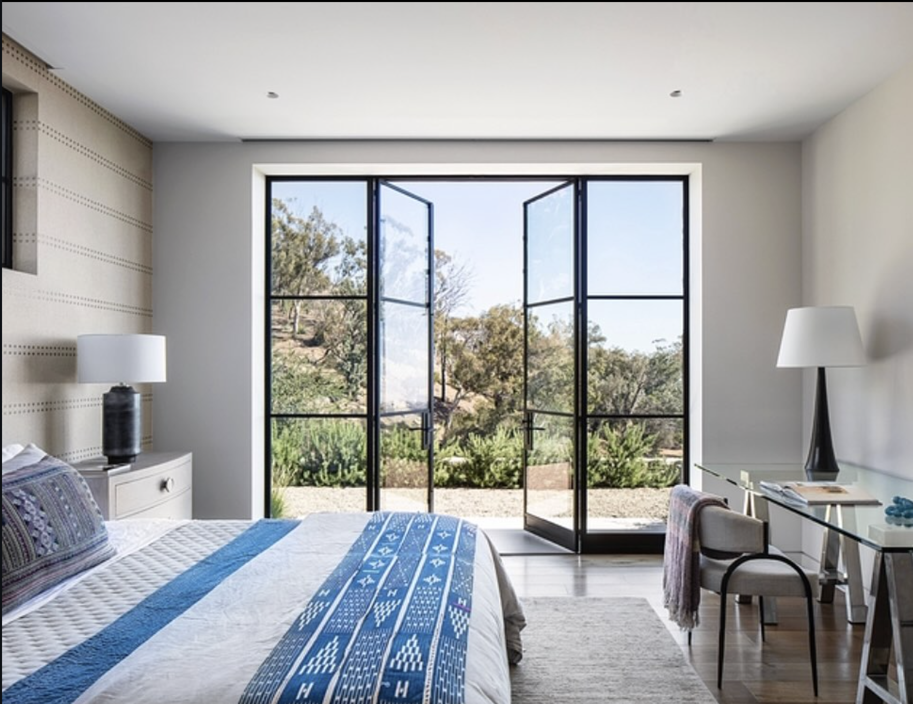 A bedroom with a large open window overlooking a garden and trees outside. The bed has a blue and white quilt and patterned pillows. To the right, there is a desk with a glass top, a chair, and a table lamp. On the left, a white nightstand with a black lamp is next to the bed. The room has light-colored walls, a beige rug, and ample natural lighting.