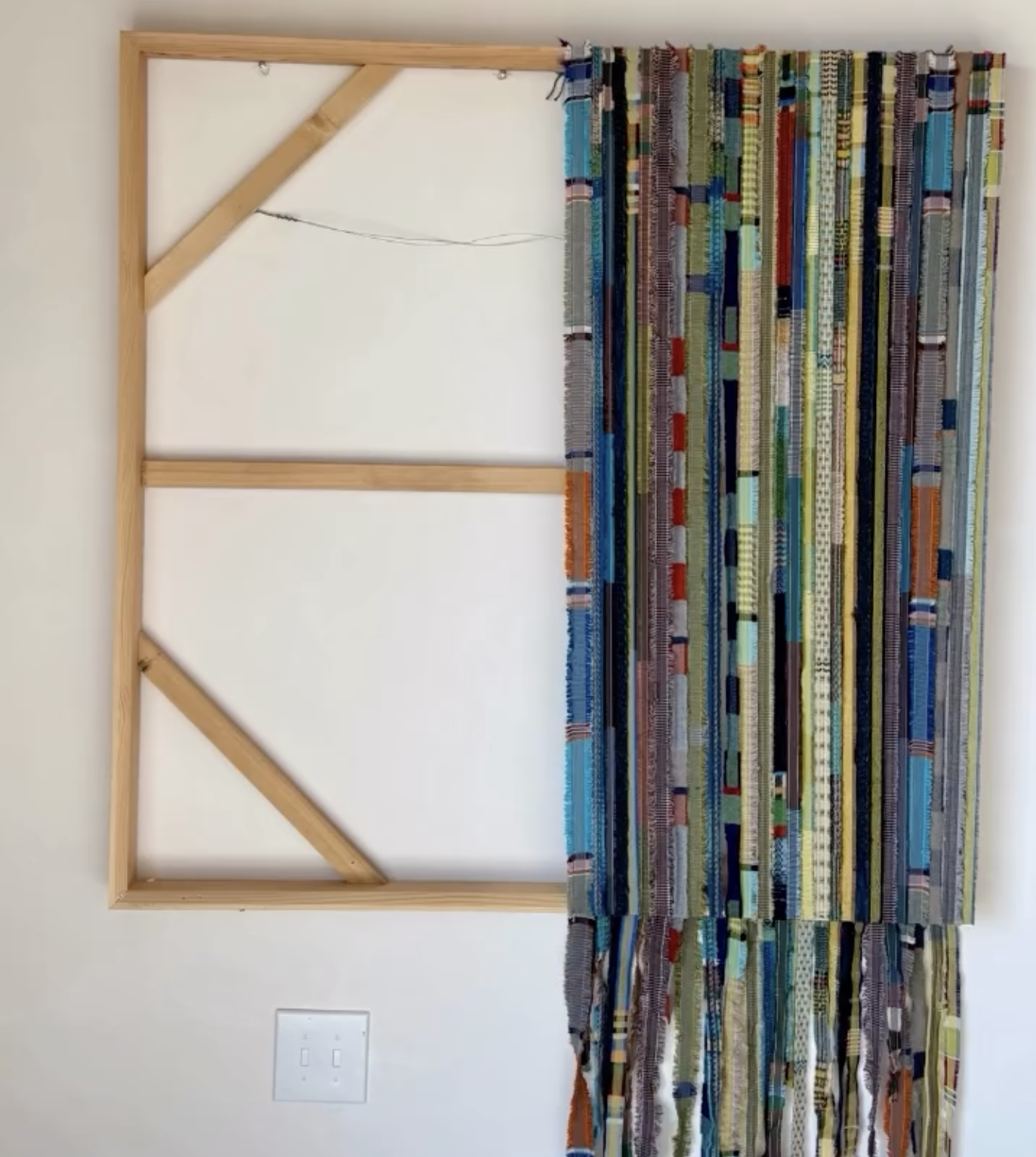 Mixed media artwork featuring a wooden geometric frame and colorful strips of fabric or paper attached underneath.