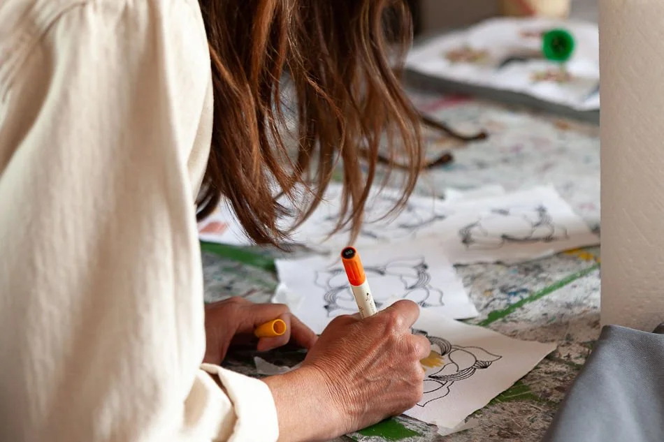 Person with curly hair drawing on paper with markers, surrounded by more drawings and art supplies on a table.