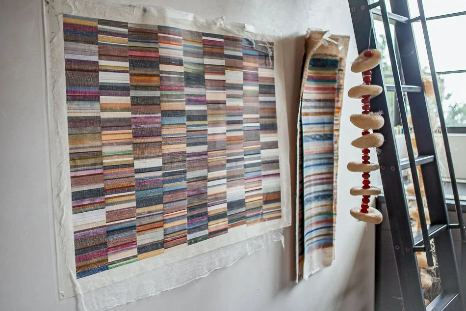 Colorful woven textile swatches and a hanging bead or shell decoration near a black ladder beside a window.