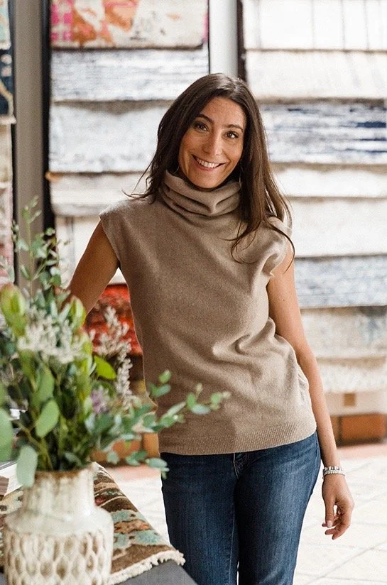 Dara Kay Barker smiling in a cozy beige sleeveless turtleneck sweater and dark jeans, standing inside a rustic decorated space with plants and wooden panels in the background.