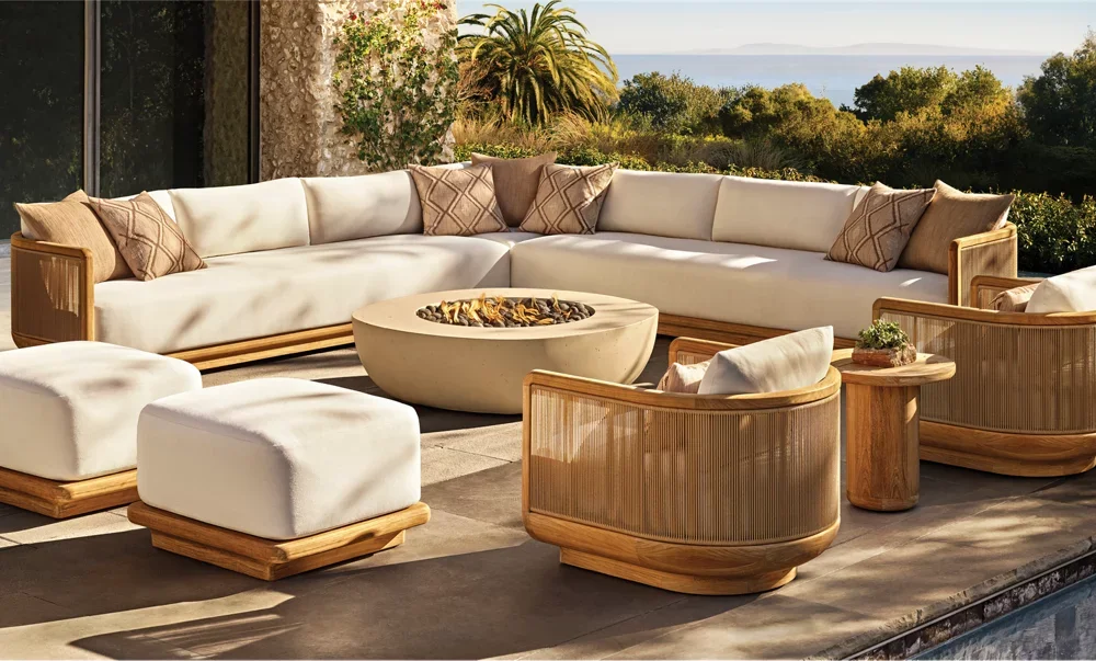 Outdoor patio with white sectional sofa, matching ottomans, and wooden armchairs around a large fire pit, with lush greenery and trees in the background and a view of the water in the distance.