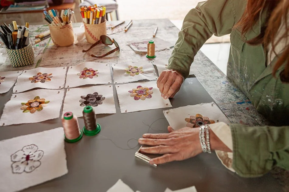 Person working on embroidery designs with floral patterns on fabric, sewing tools, spools of thread, and markers on a work table.