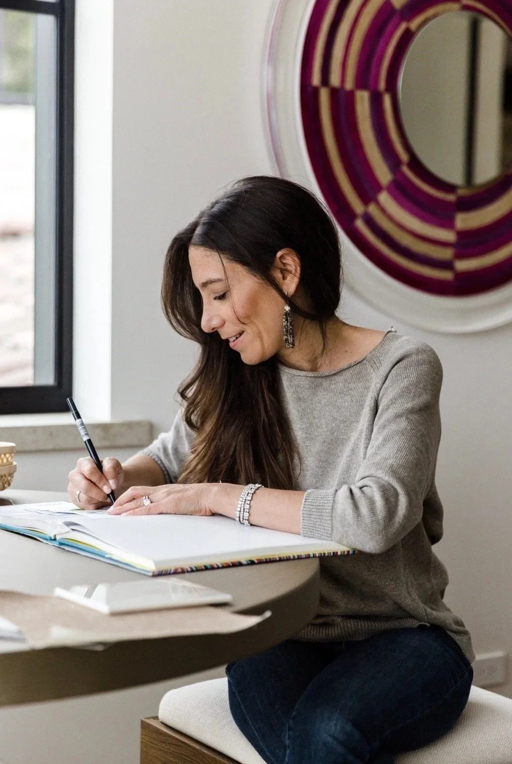 Dara Kay Barker sitting at a desk, writing in a notebook with a pen, in a well-lit room with a large circular decorative mirror on the wall.