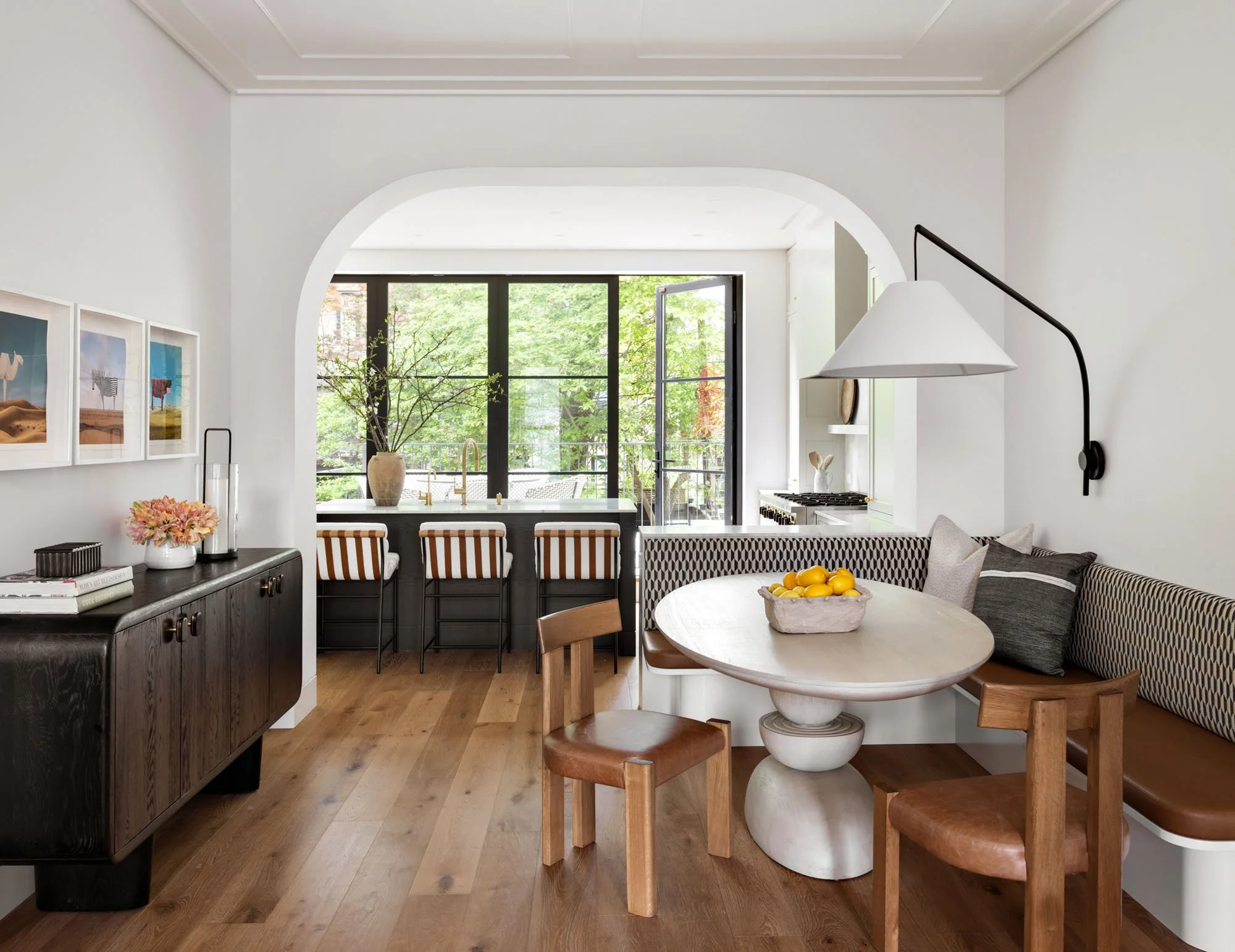 A bright, modern kitchen and dining area with large windows and greenery outside. The kitchen features a window with a view of trees, a vase with branches, and a black sideboard with books and flowers. The dining area has a round marble table with wooden chairs and lemons in a bowl, with barstools and buffet in the background.