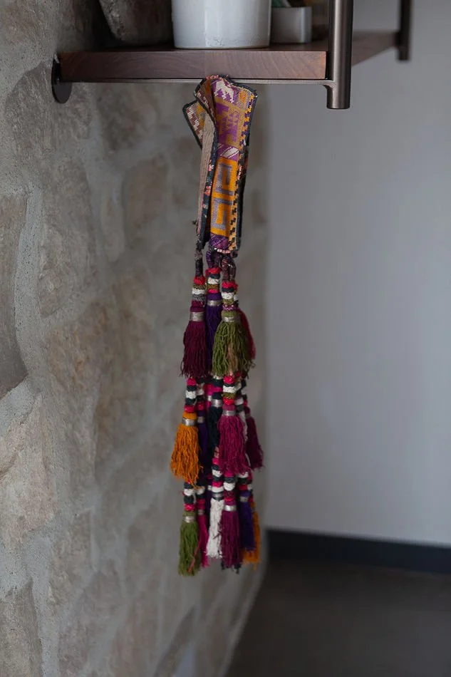 Colorful woven lanyard with tassels hanging from a metal hook on a wooden shelf, against a stone wall.