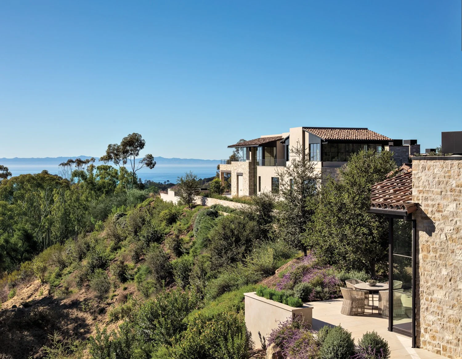 Luxury modern house on a hillside with lush green trees, overlooking a body of water and distant mountains under a clear blue sky.