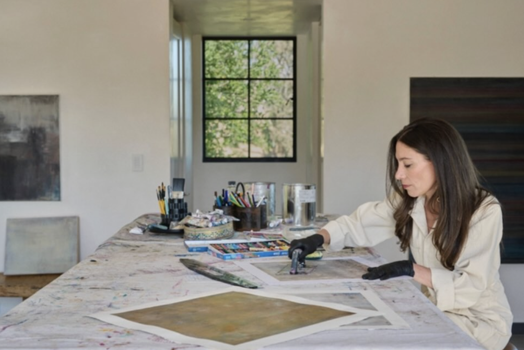 Dara Kay Barker working on art projects at a large table in a bright room with a window showing green trees outside. The table is covered with art supplies, including markers, paint, and papers.