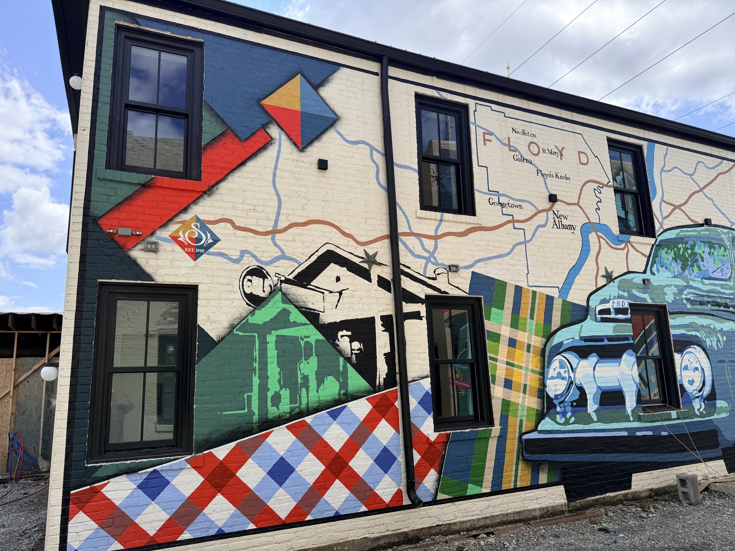 A mural painted on the side of a brick building depicts various colorful symbols and images, including a fire symbol, a vintage fire truck, and a map of a local area with roads and neighborhood names.