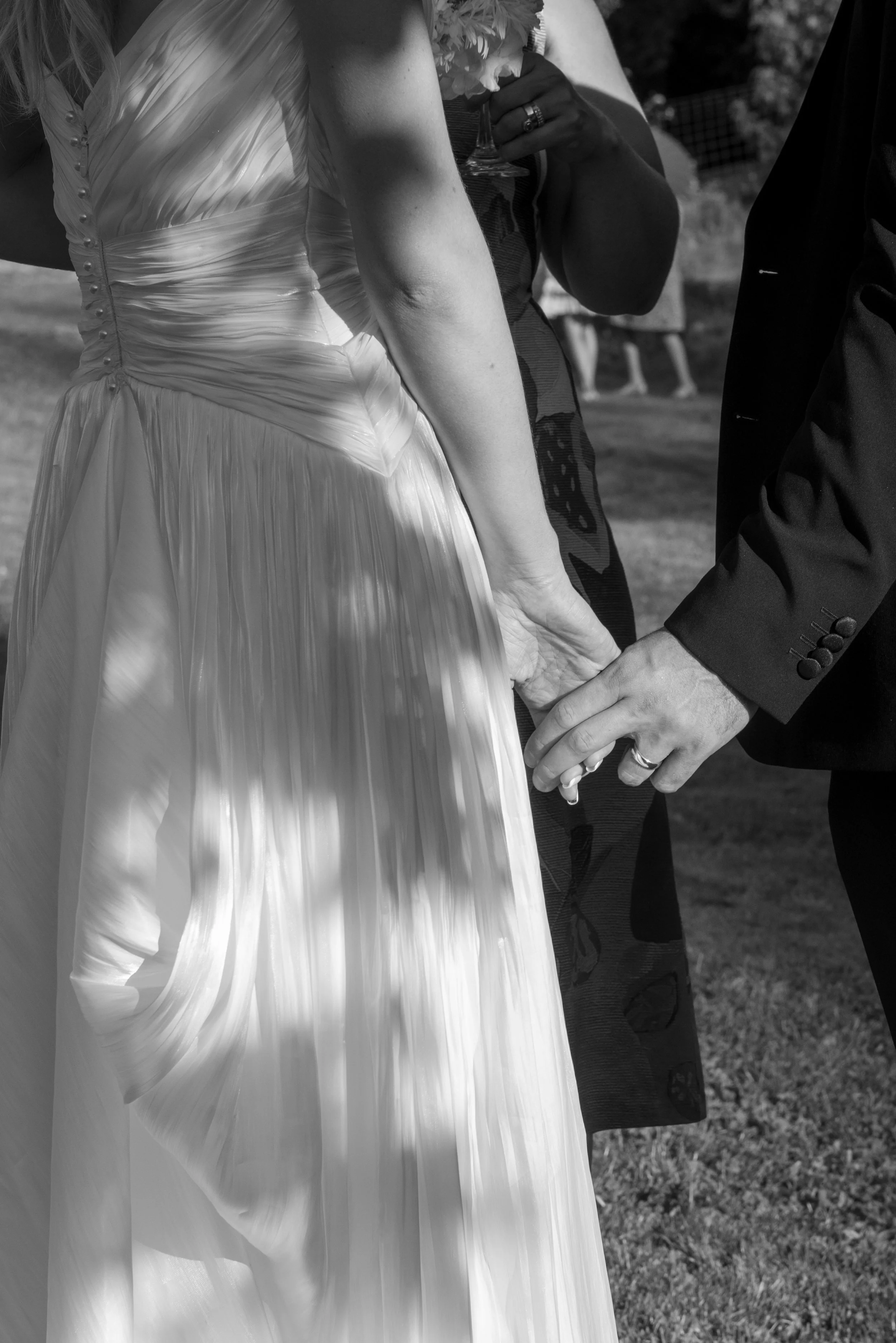 Close-up of a wedding ceremony where a bride and groom are holding hands, with the focus on their hands and a wedding ring on the groom's finger. The bride is wearing a satin wedding gown and the groom is in a dark suit, holding a wedding ring.