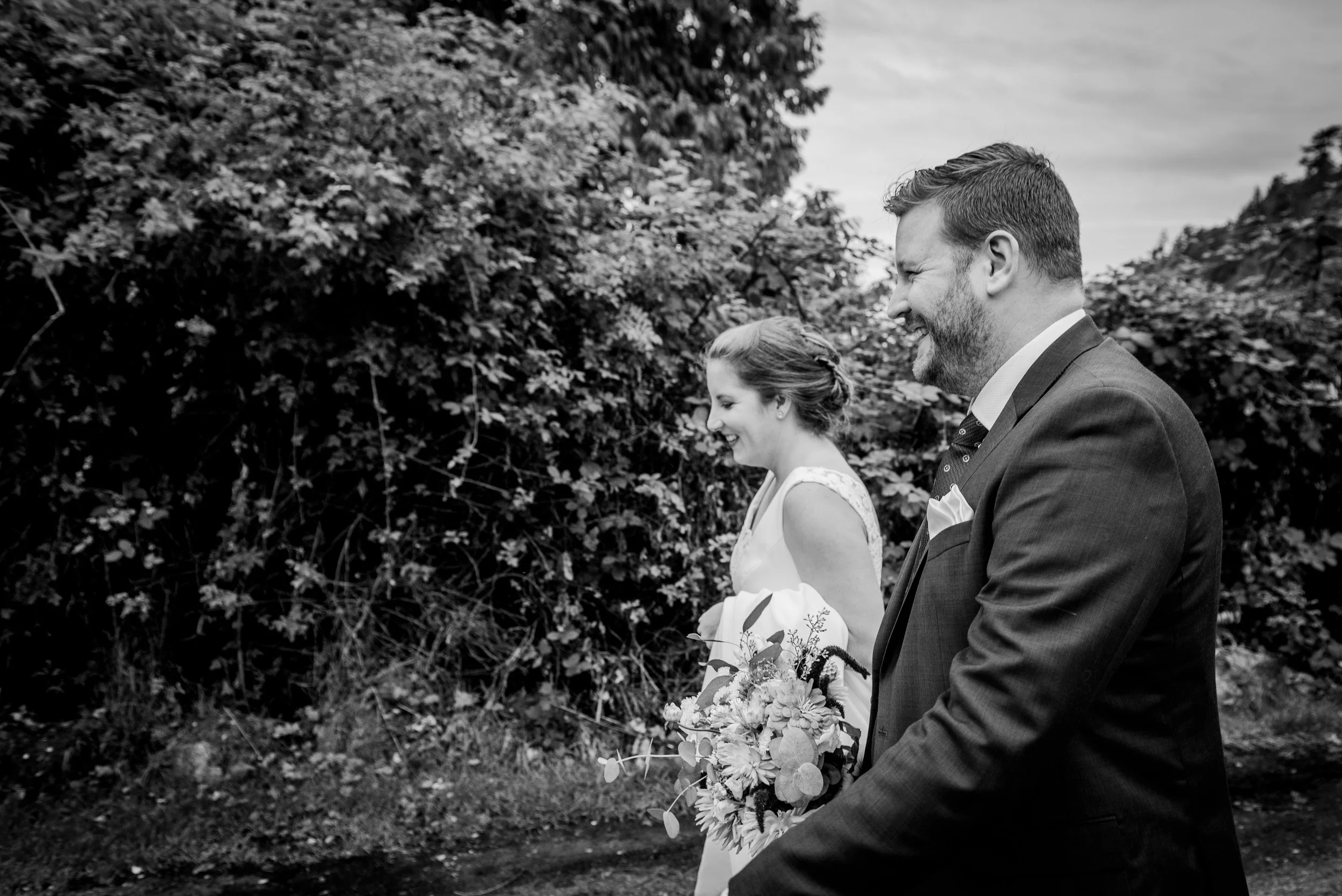 A black and white photo of a bride and groom walking outdoors, smiling and holding flowers, with trees and shrubbery in the background.