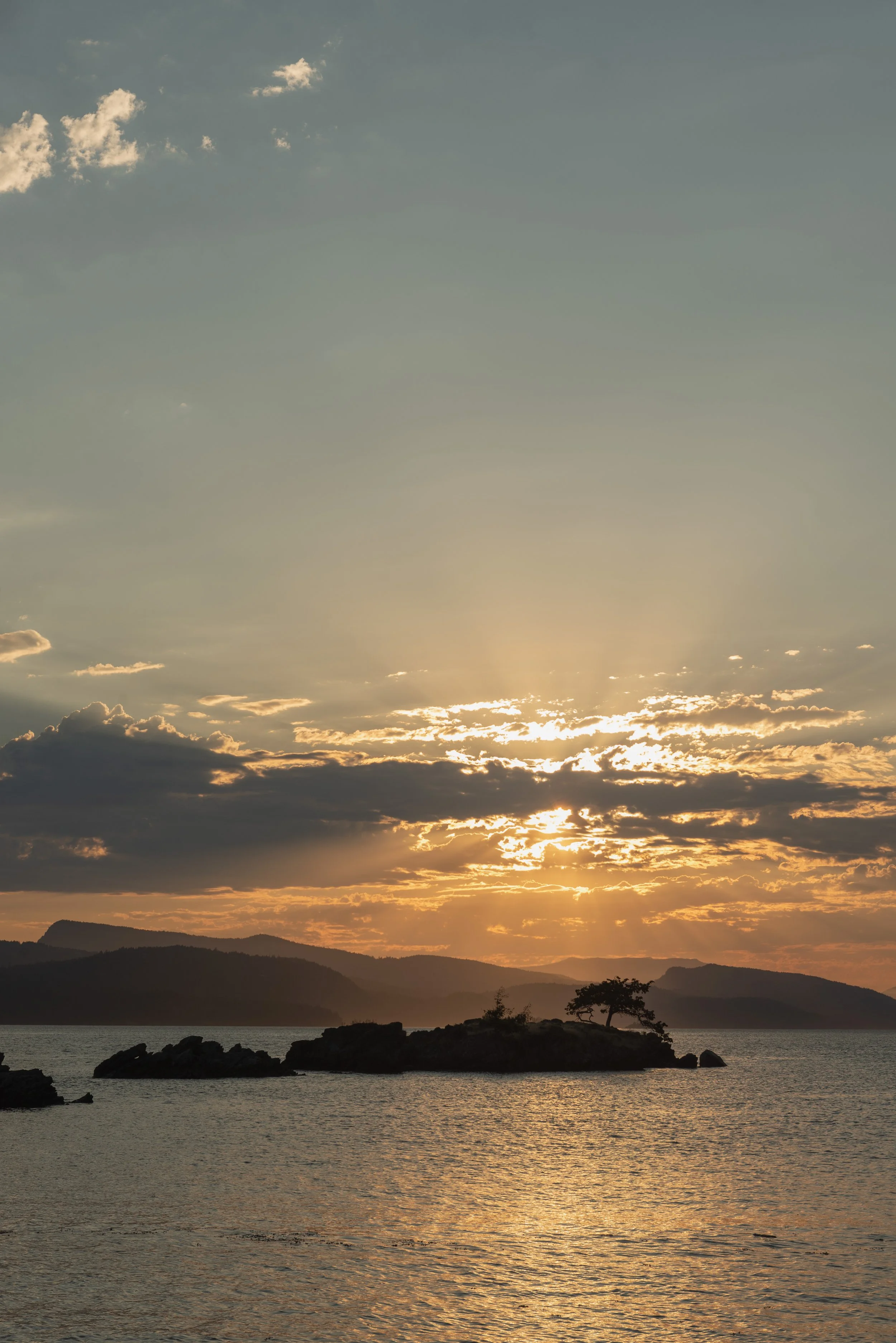 A sunset over a body of water with mountains and trees silhouetted in the foreground, and clouds illuminated by the setting sun in the sky.