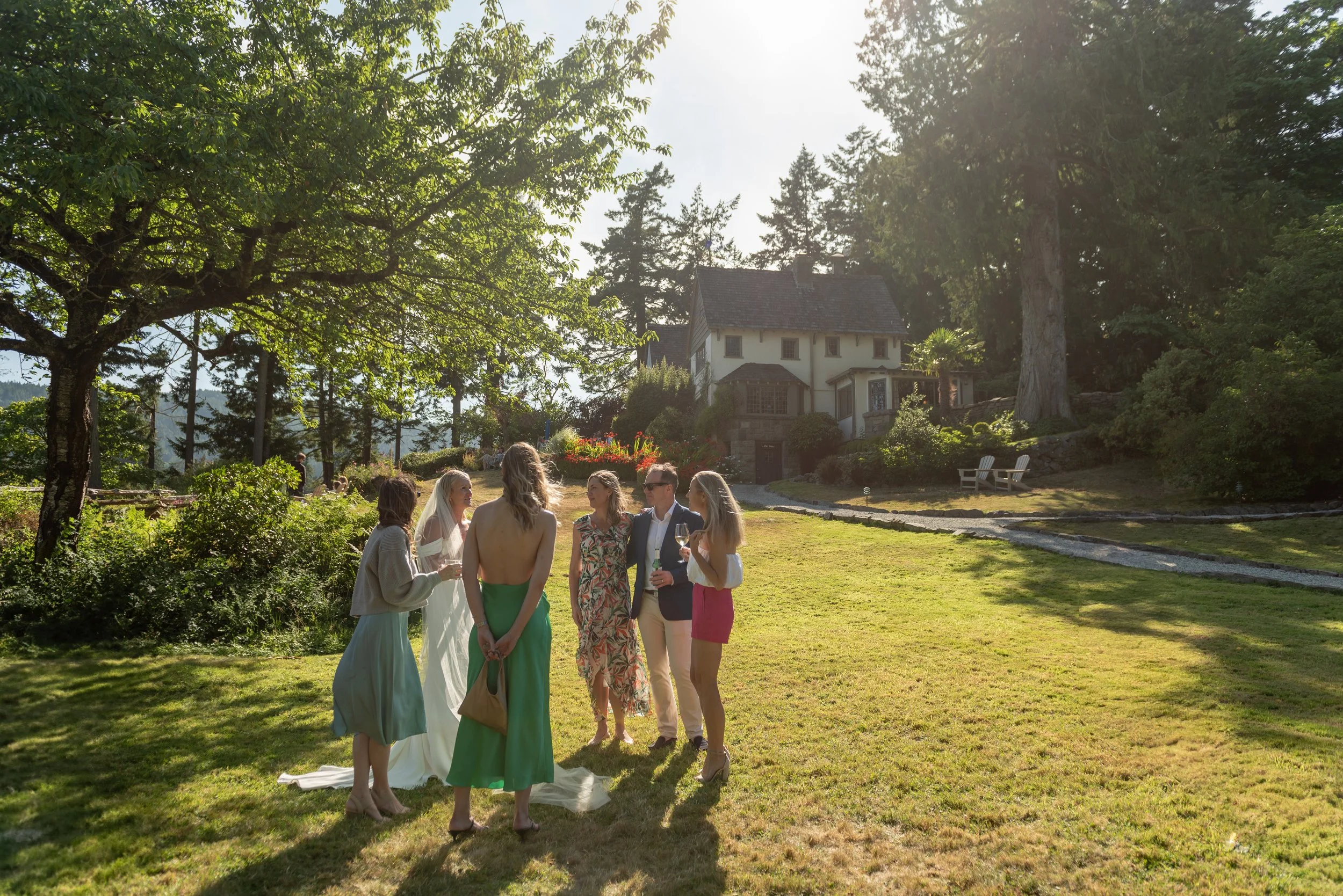 Group of people at an outdoor wedding, standing on grass near a house surrounded by trees, with sunlight filtering through the leaves.