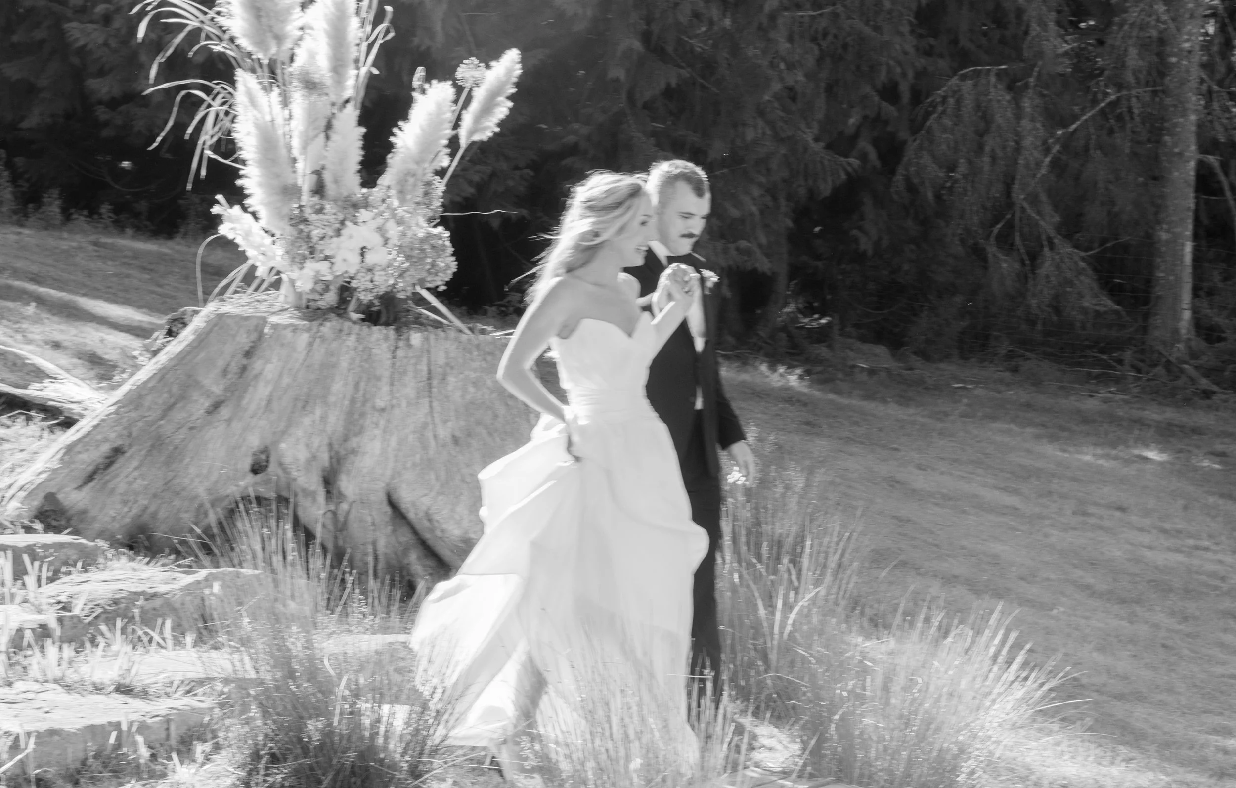 A bride and groom walking outdoors past a large tree trunk with floral decorations in a natural setting.