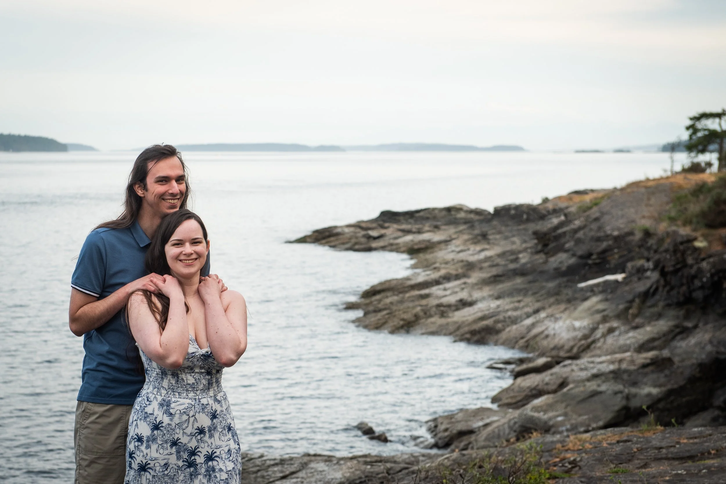 A man and woman smiling near a rocky shoreline with water and islands in the background.