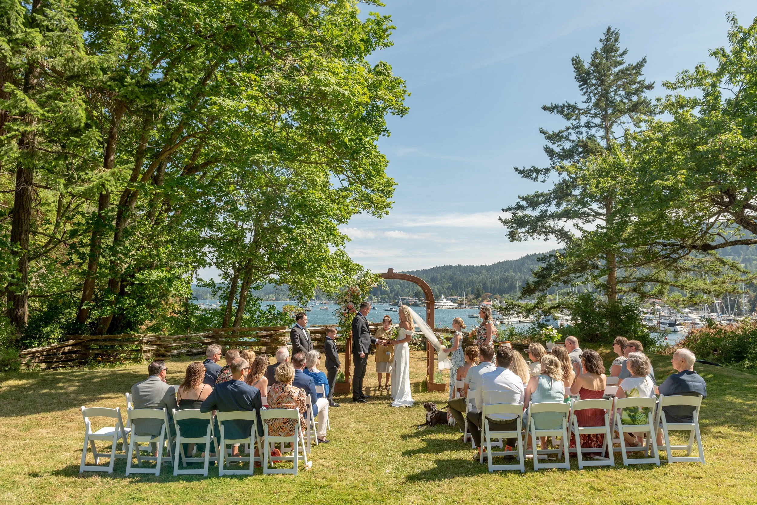 Outdoor wedding ceremony with as guests seated on white chairs, a bride and groom exchanging vows under an arch, trees and a marina with boats in the background.