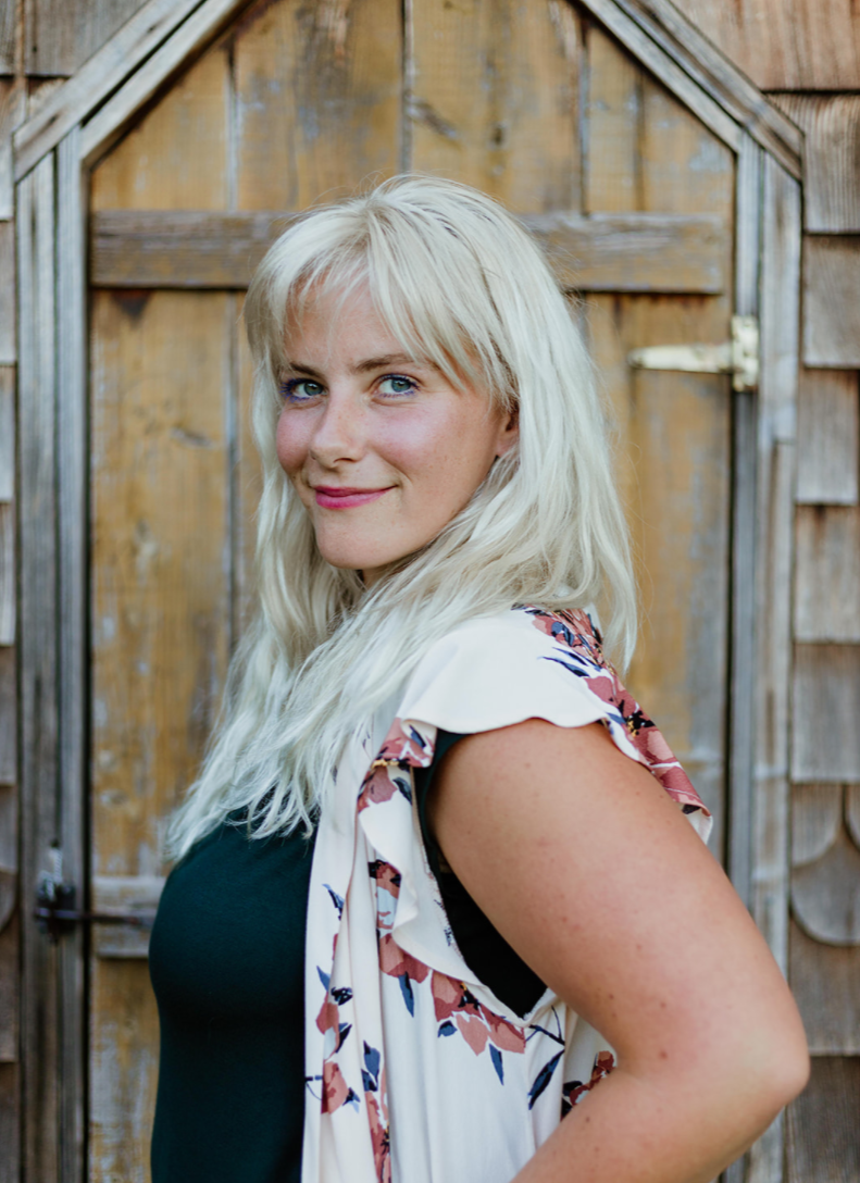 A woman with long, wavy blonde hair and blue eyes, smiling while standing outdoors in front of a wooden fence or door.