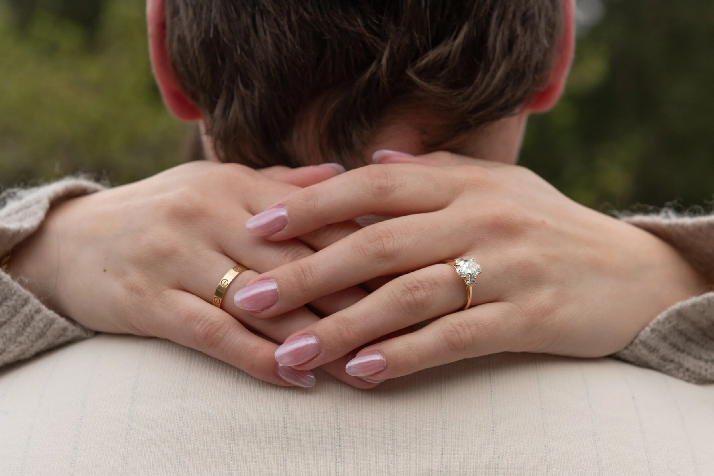 Close-up of a person with short brown hair, with their face obscured, resting their head on their crossed arms. The person’s hands are visible with rings, including a diamond ring on the ring finger. The background is blurred greenery.
