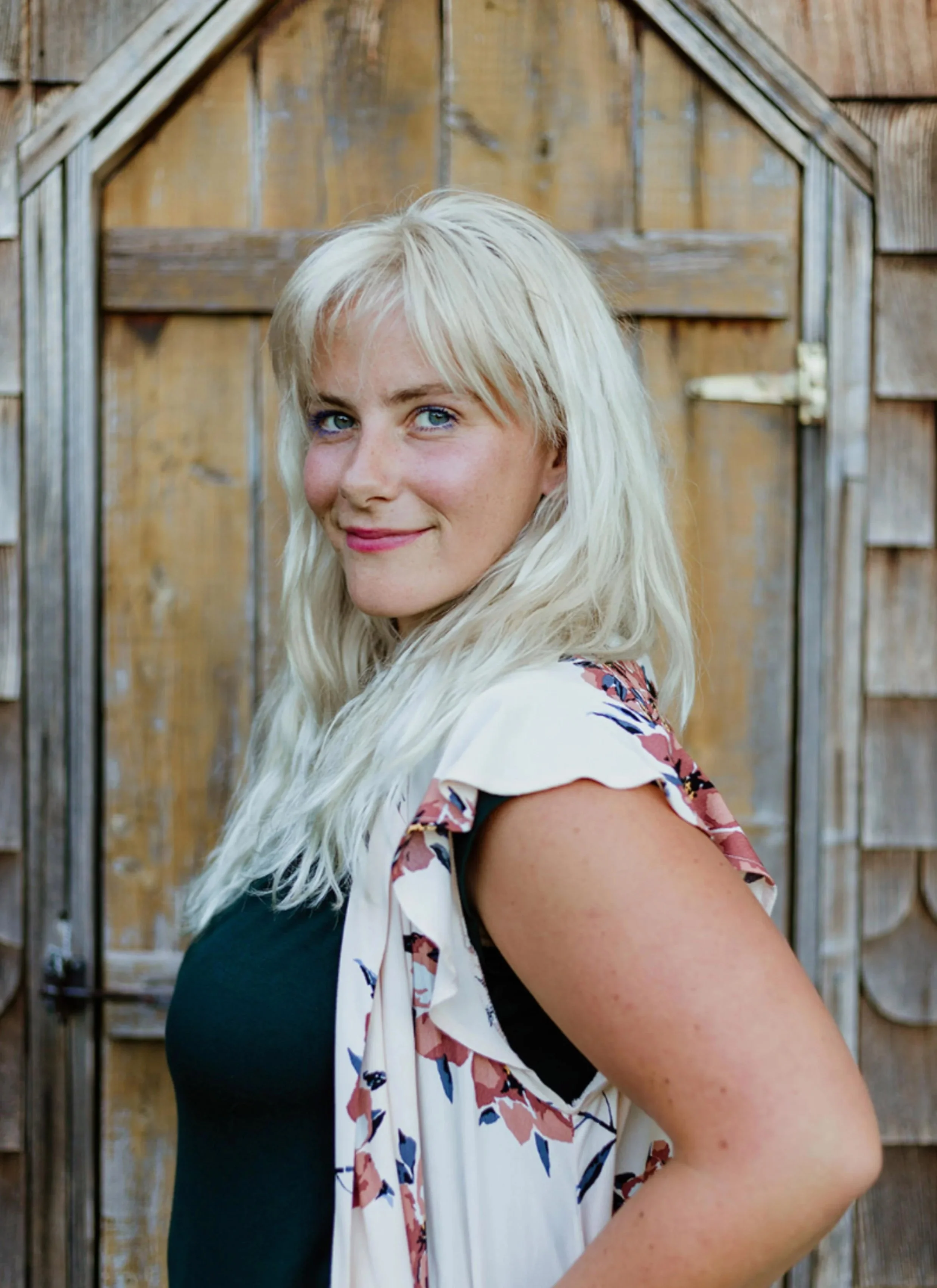 A woman with long, wavy blonde hair and blue eyes standing outdoors in front of a wooden fence, smiling slightly and looking at the camera.