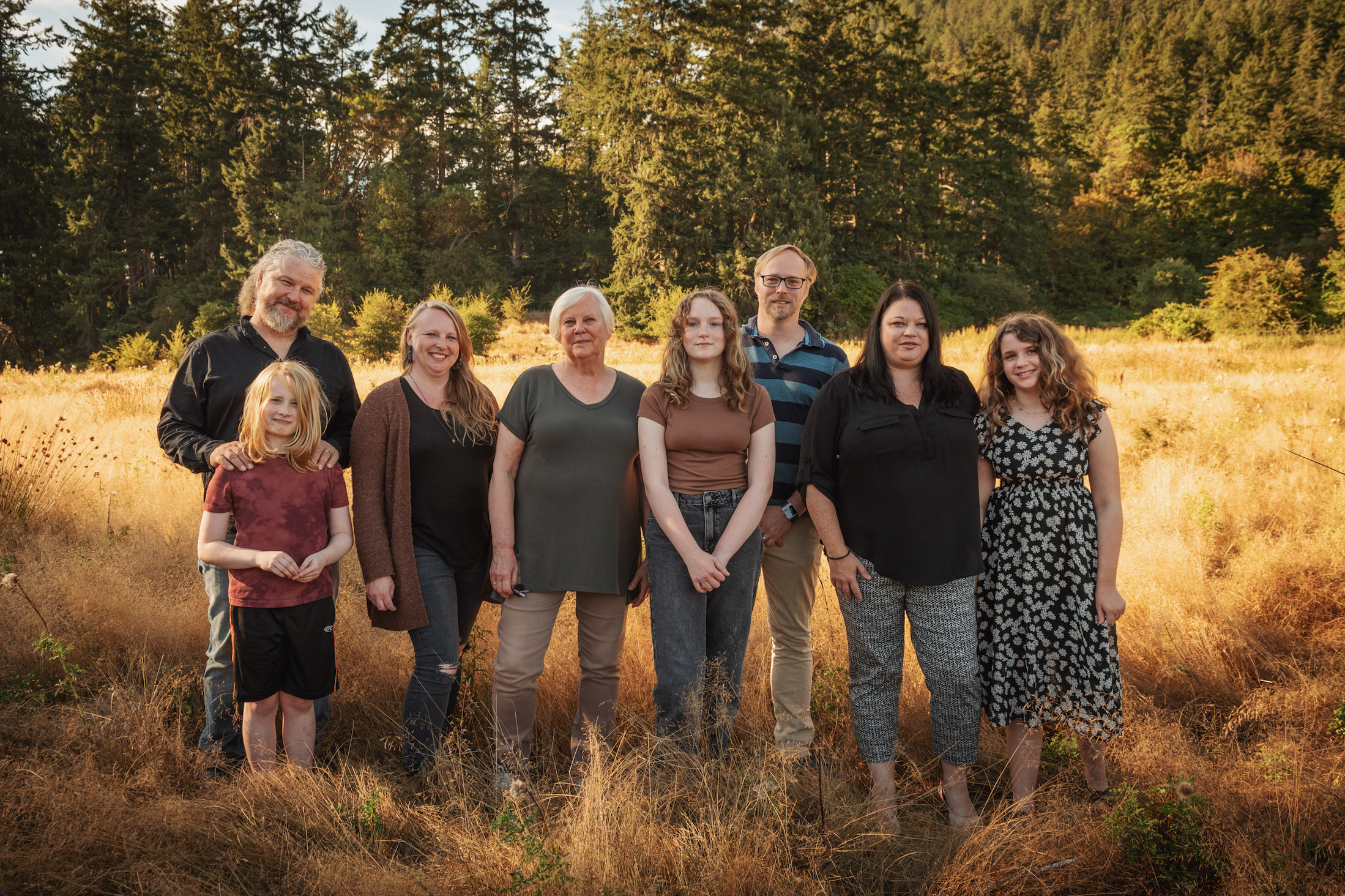 Family of eight poses outdoors in a field with tall trees in the background during sunset.