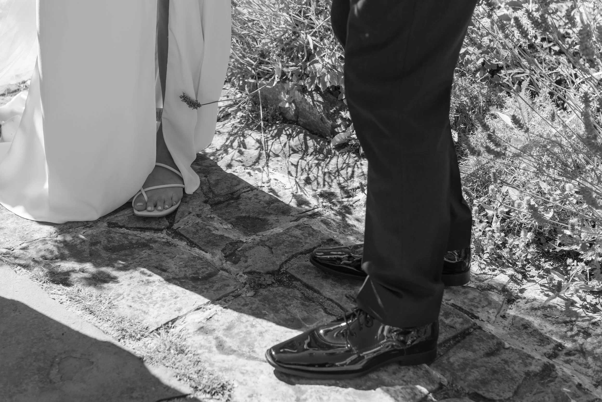Close-up of a bride and groom standing outdoors on a rocky surface. The bride is wearing a white dress and sandals, visible from the knee down. The groom is dressed in dark pants and shiny black shoes, also visible from the knee down. Vegetation is i