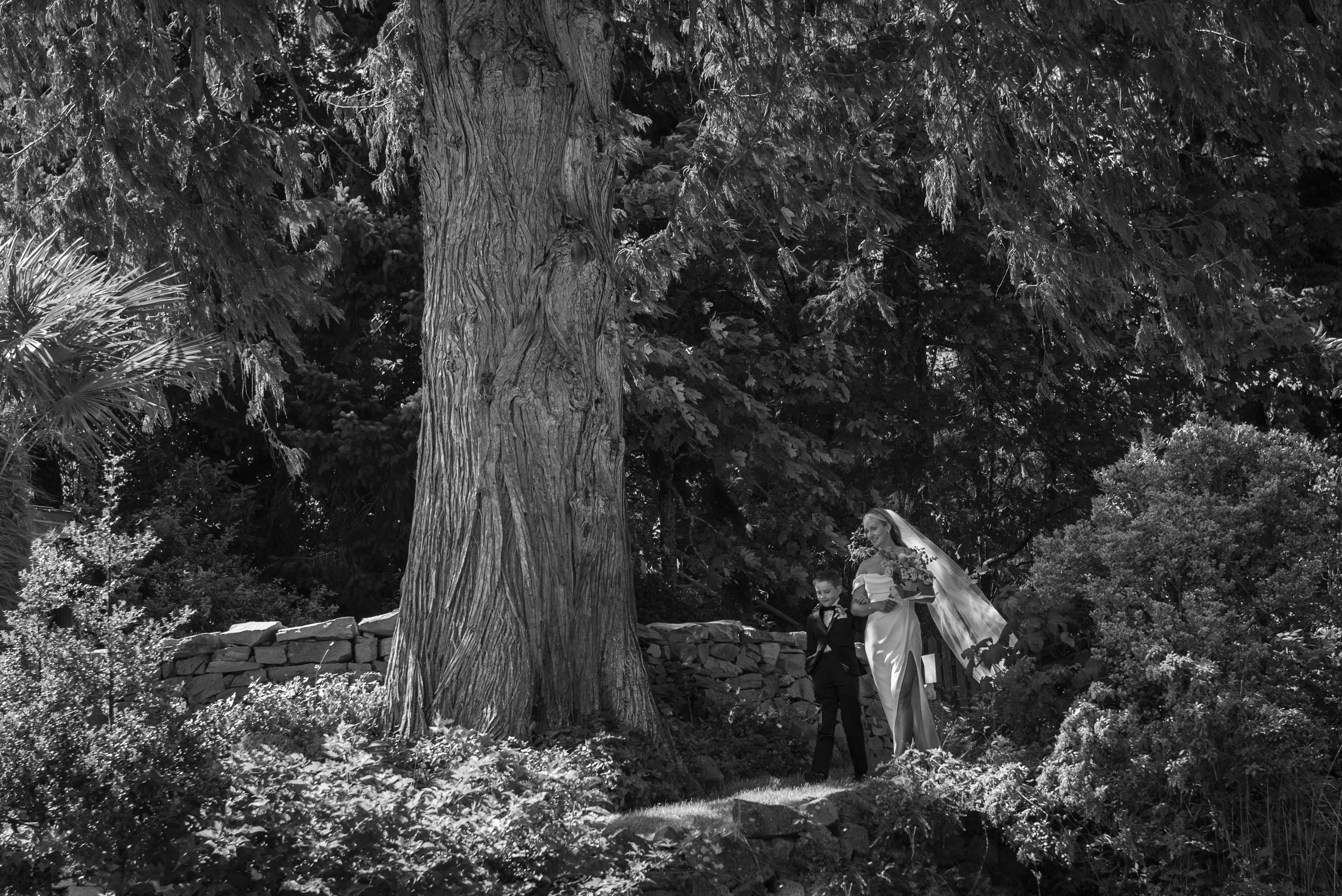 A black-and-white photo of a bride and a young boy walking through a wooded outdoor area during a wedding. The bride is in a long dress with a veil, and the boy is dressed in a suit.
