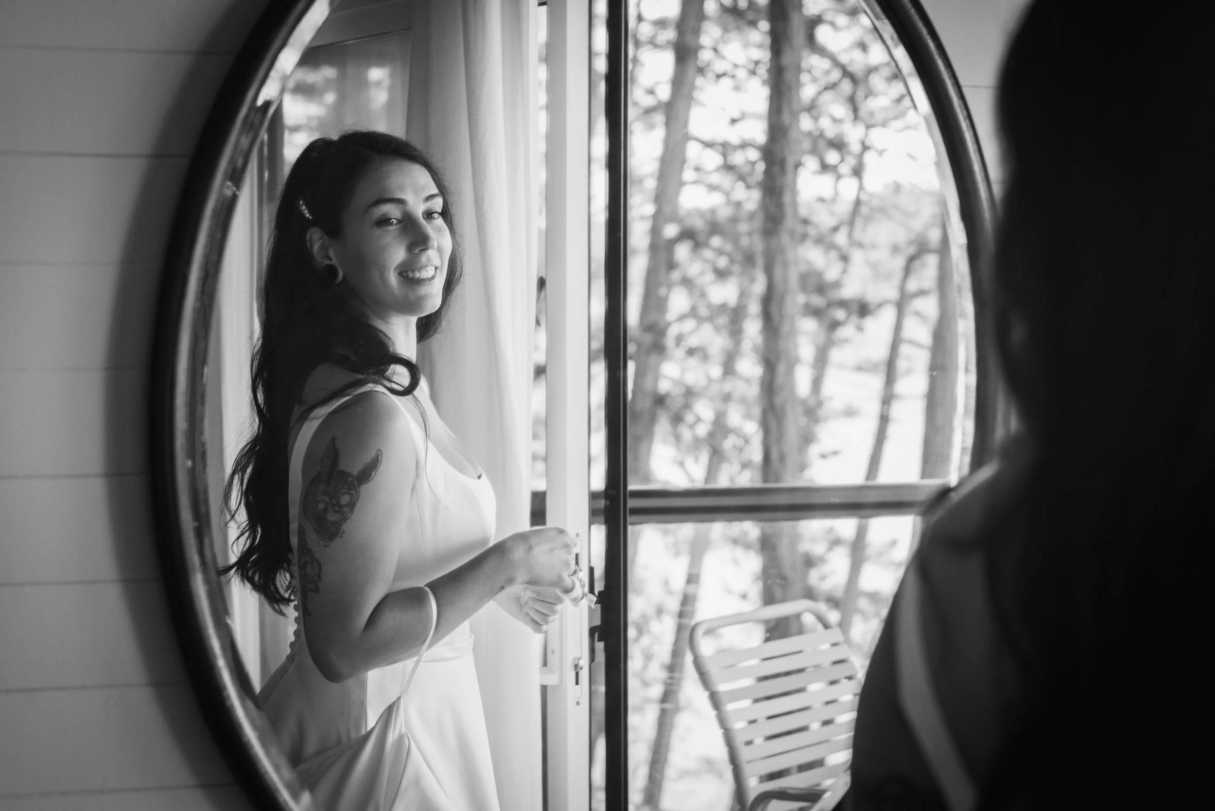 Black and white photo of a woman with tattoos looking into a mirror inside a trailer or camper, with trees visible outside through an open window.