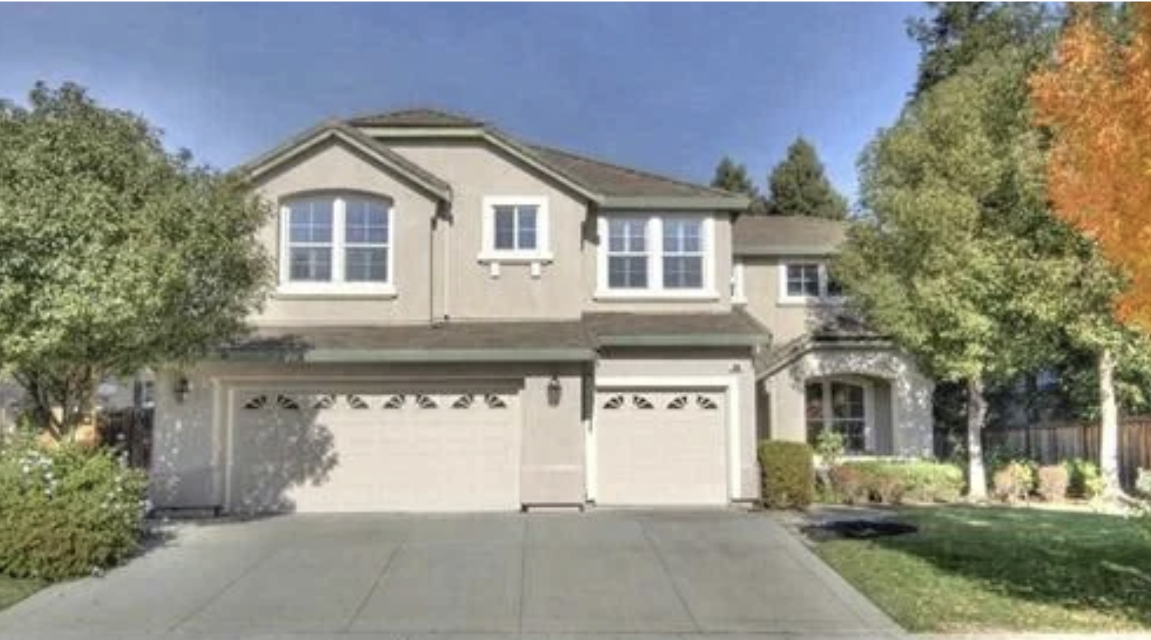 A two-story suburban house with a two-car garage, beige exterior, and white trim, surrounded by green trees and a small front yard with a driveway.