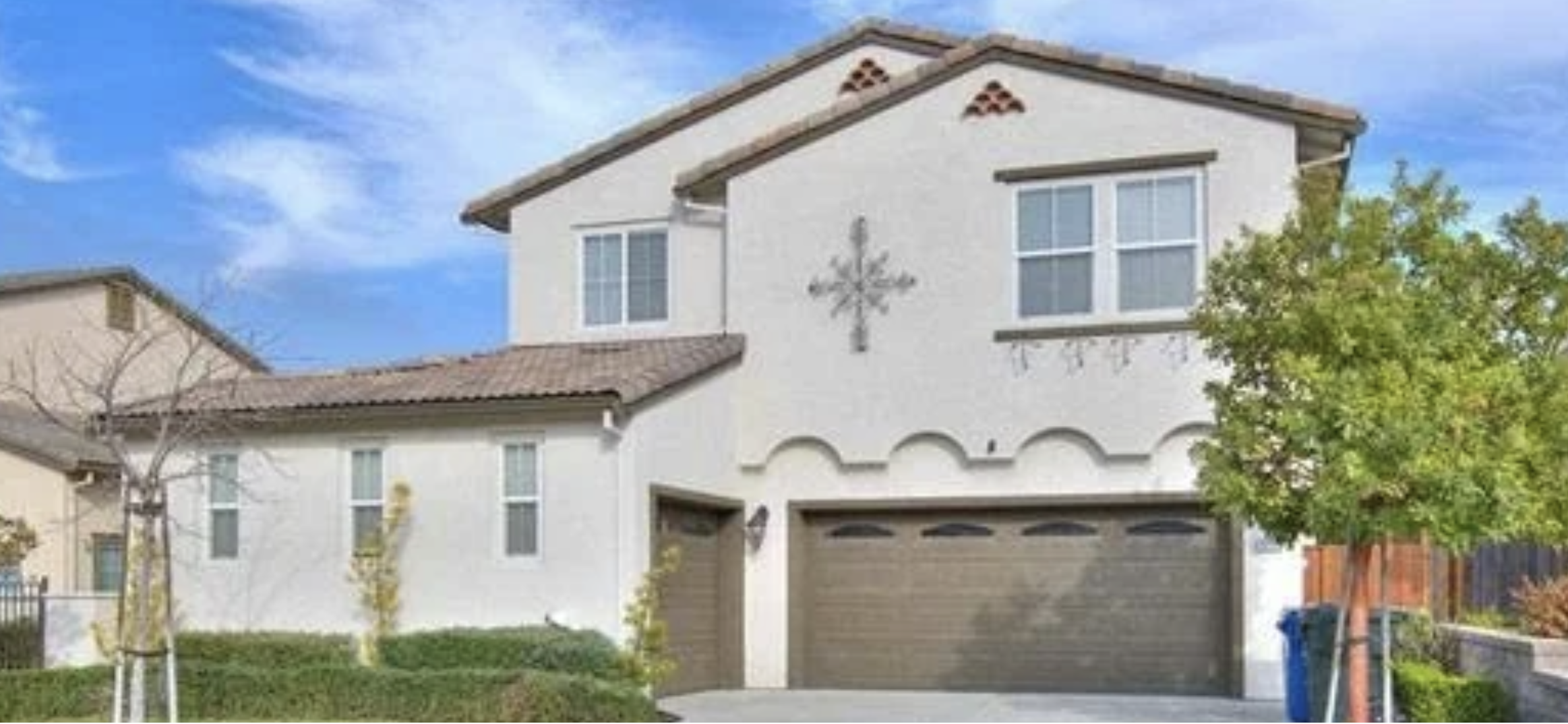 Front view of a two-story house with a decorated cross on the wall, a double garage door, and a driveway, surrounded by small trees and bushes, under a partly cloudy sky.