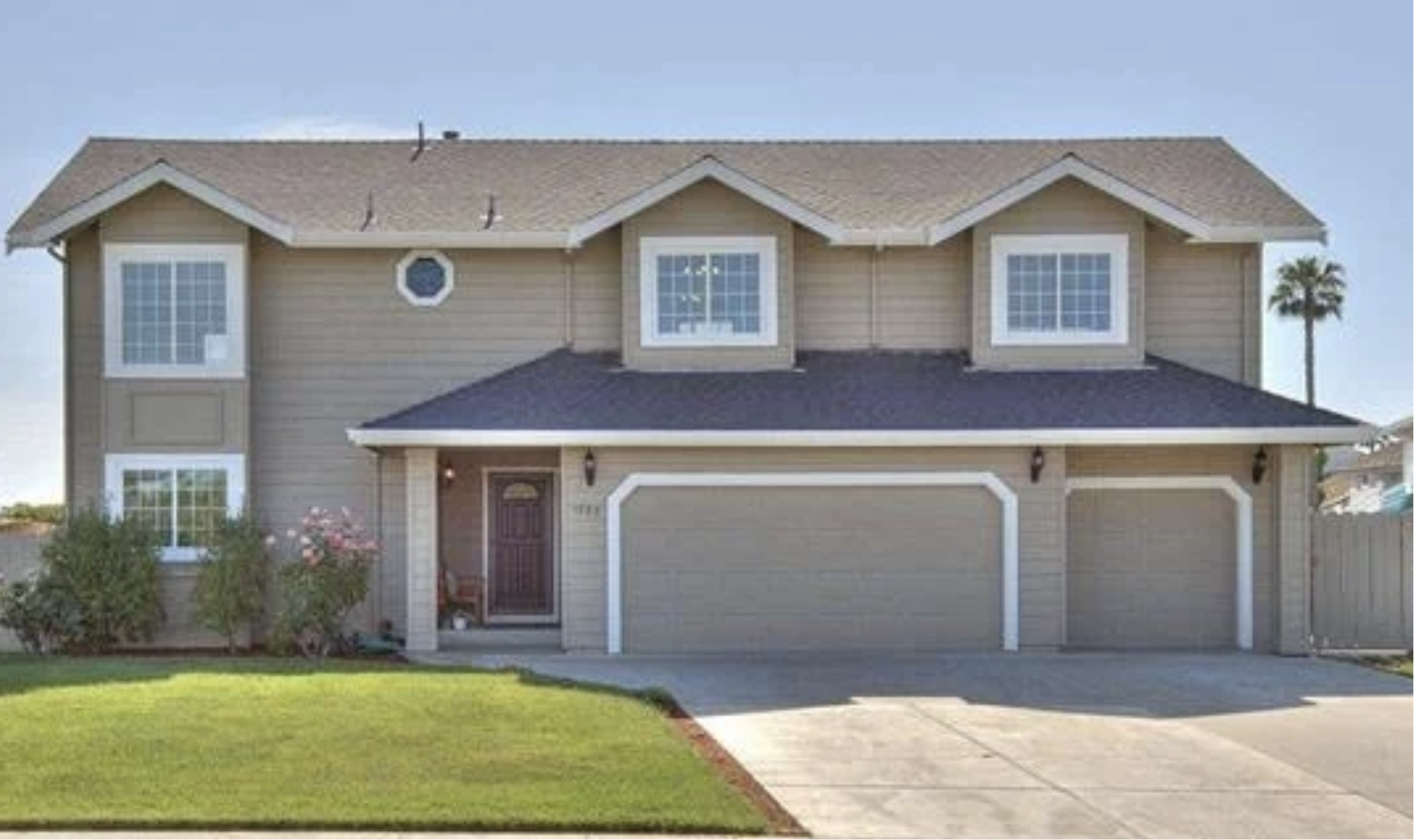 Front view of a two-story house with a dark gray garage door, beige siding, and white trim, with a lawn and sidewalk in the foreground, a bush and pink flowers near the entrance, and a palm tree to the right.